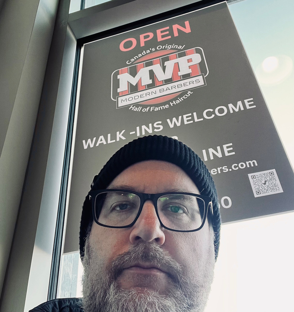 A person wearing glasses and a beanie is sitting in front of a window with a sign for MVP Modern Barbers.