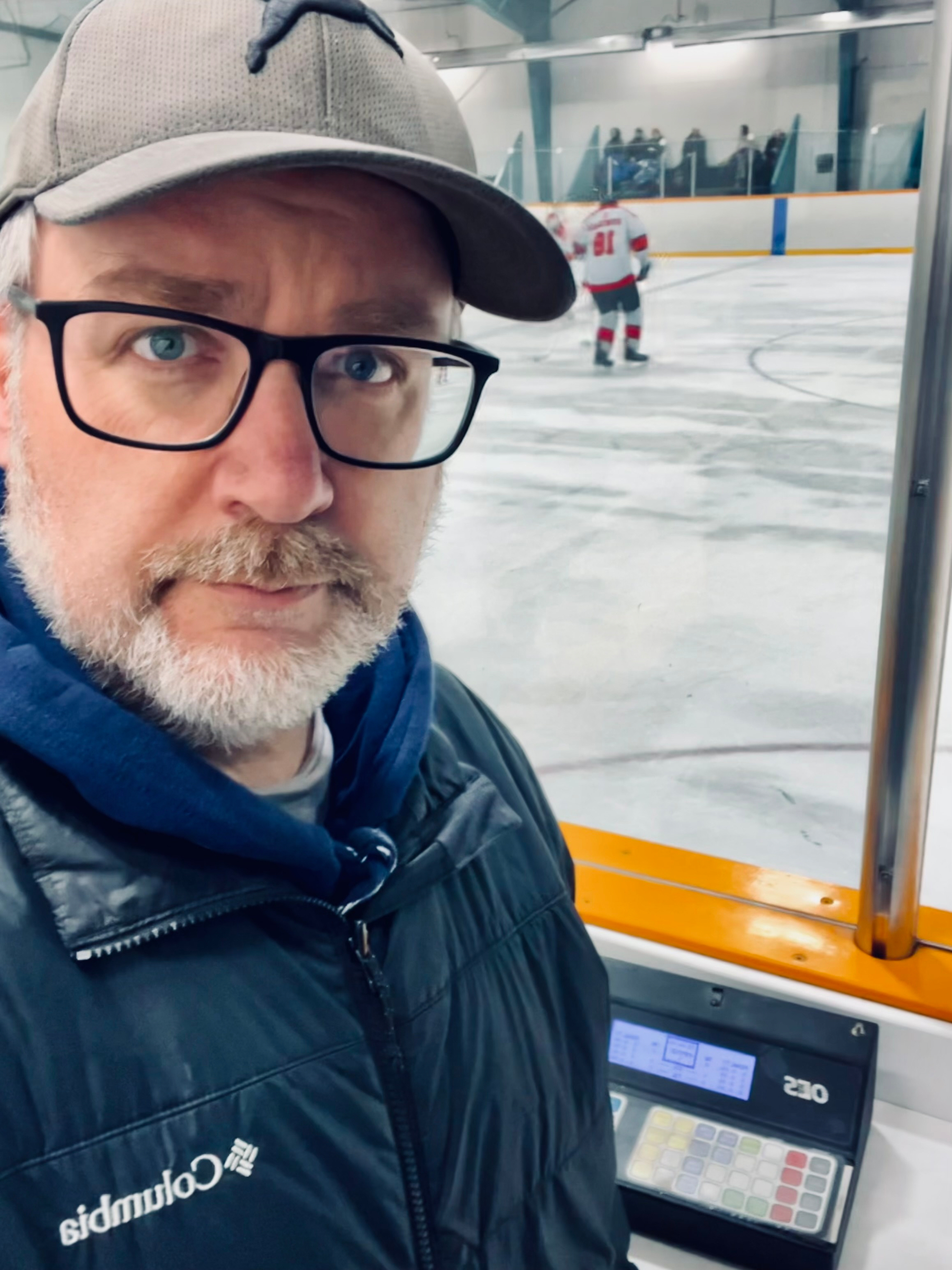 A man wearing glasses and a cap stands indoors, with a hockey game taking place in the background.
