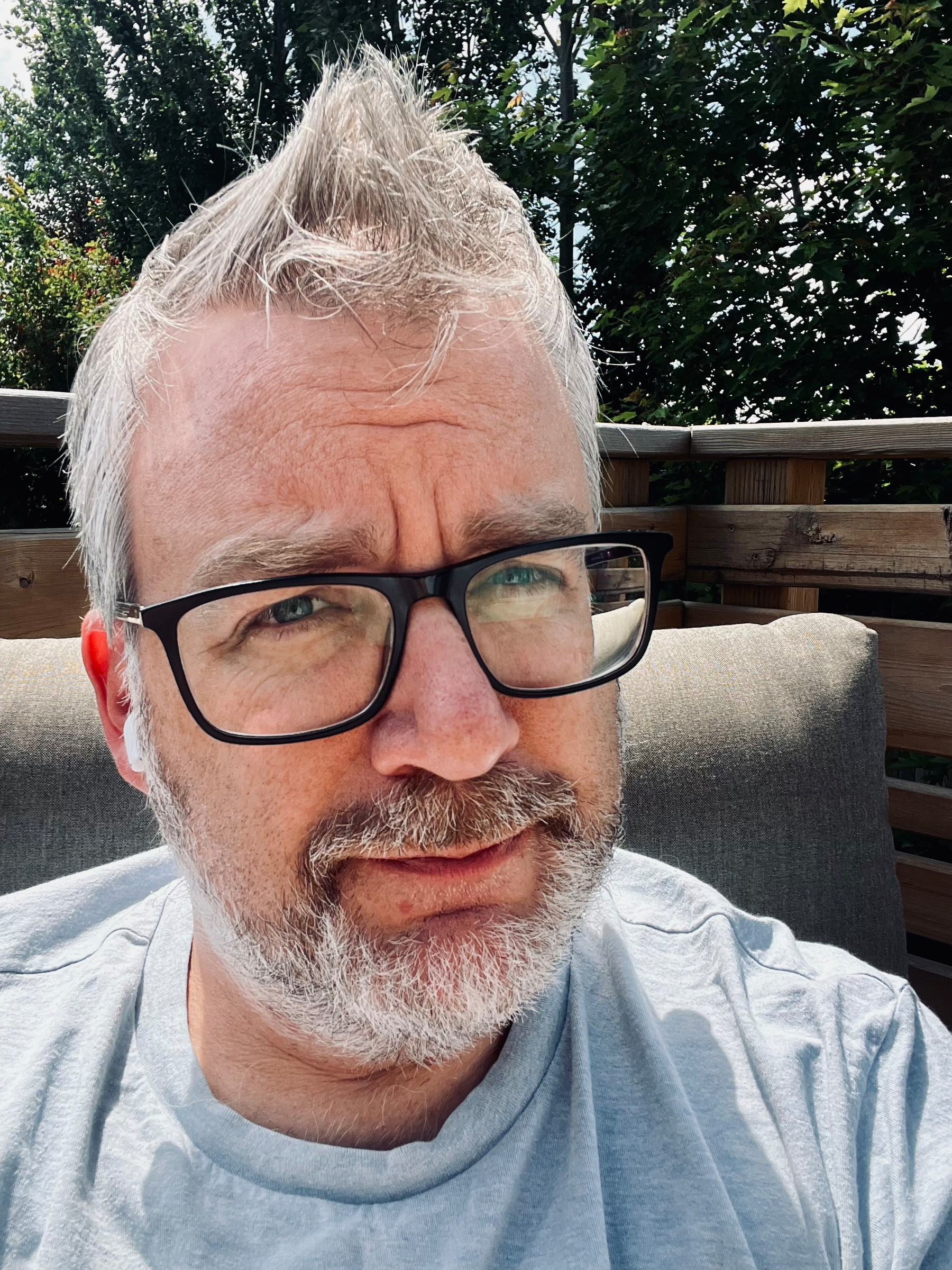 A man with gray hair, glasses, and a beard is sitting outdoors on a cushioned chair with greenery in the background.