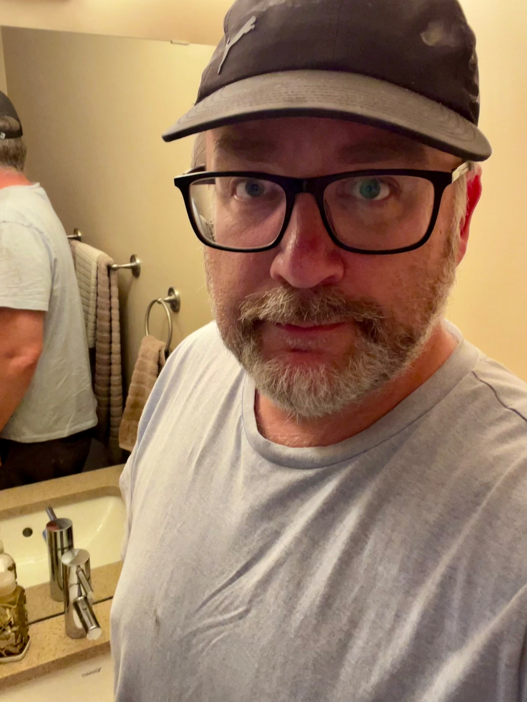 A man wearing glasses and a cap takes a selfie in a bathroom, with a reflection visible in the mirror behind him.