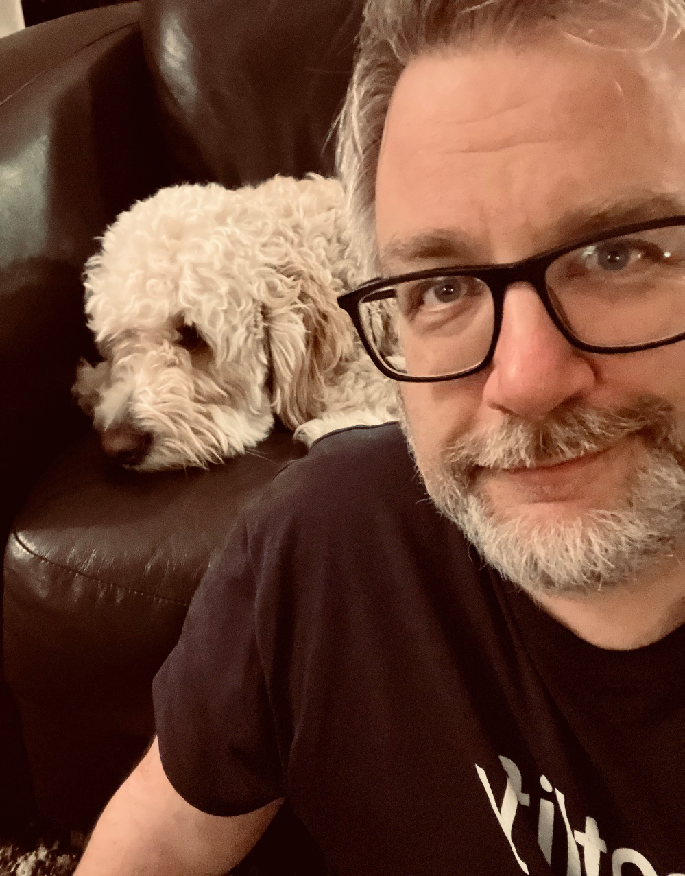A man with glasses is smiling while a fluffy white dog rests on a dark sofa behind him.