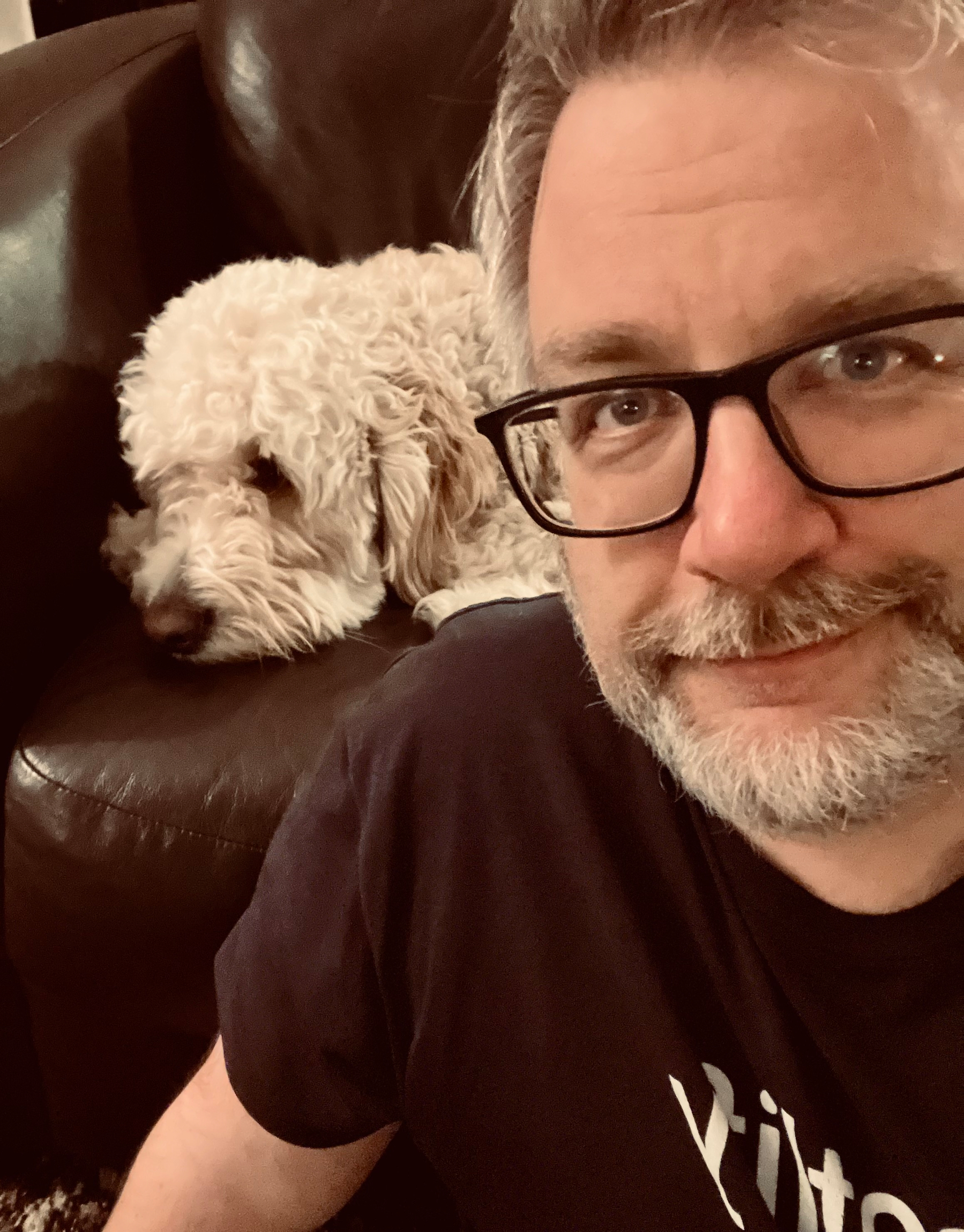A man with glasses is smiling while a fluffy white dog rests on a dark sofa behind him.