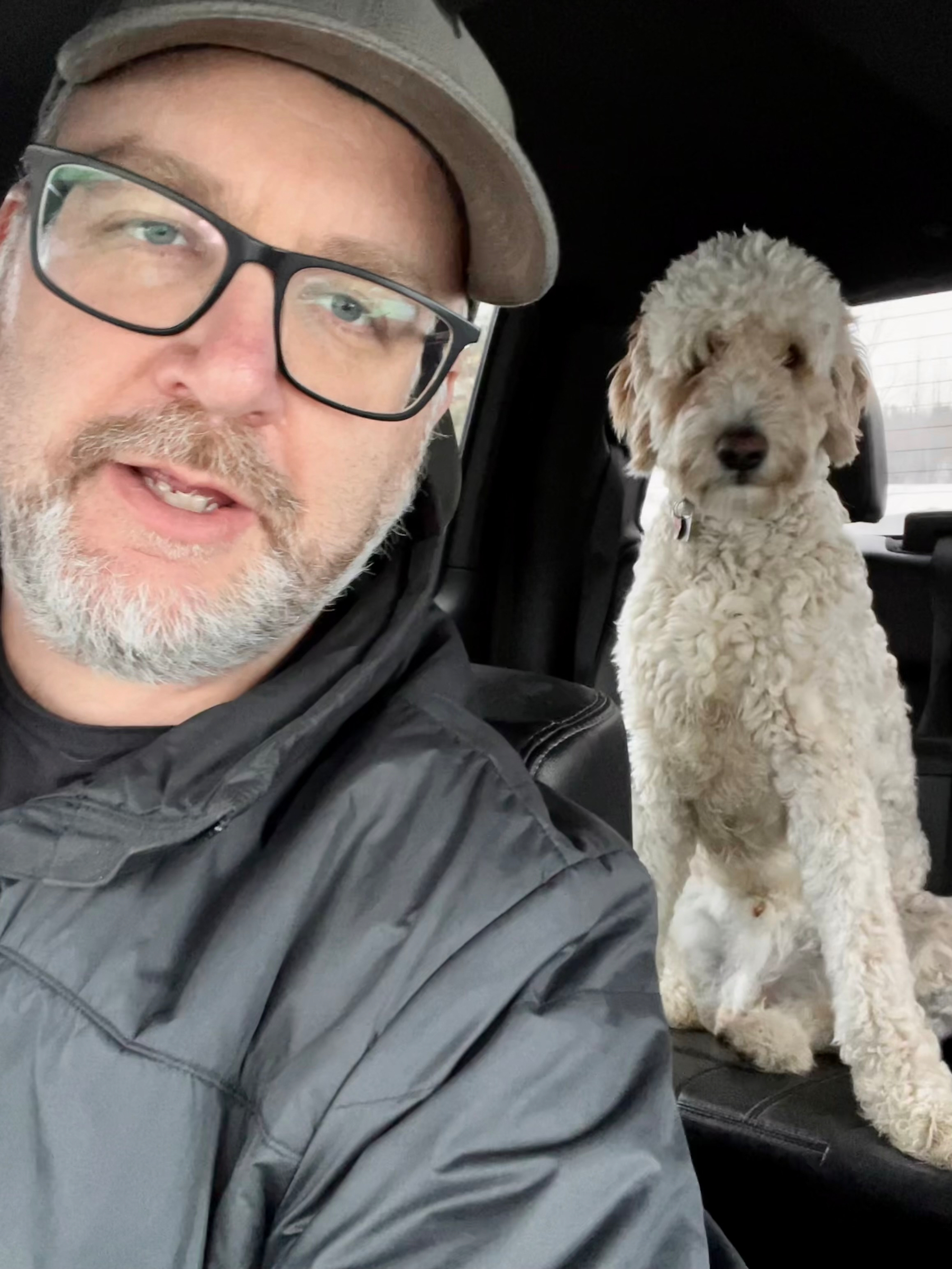 A man wearing glasses and a cap is sitting in a car with a fluffy dog in the back seat.