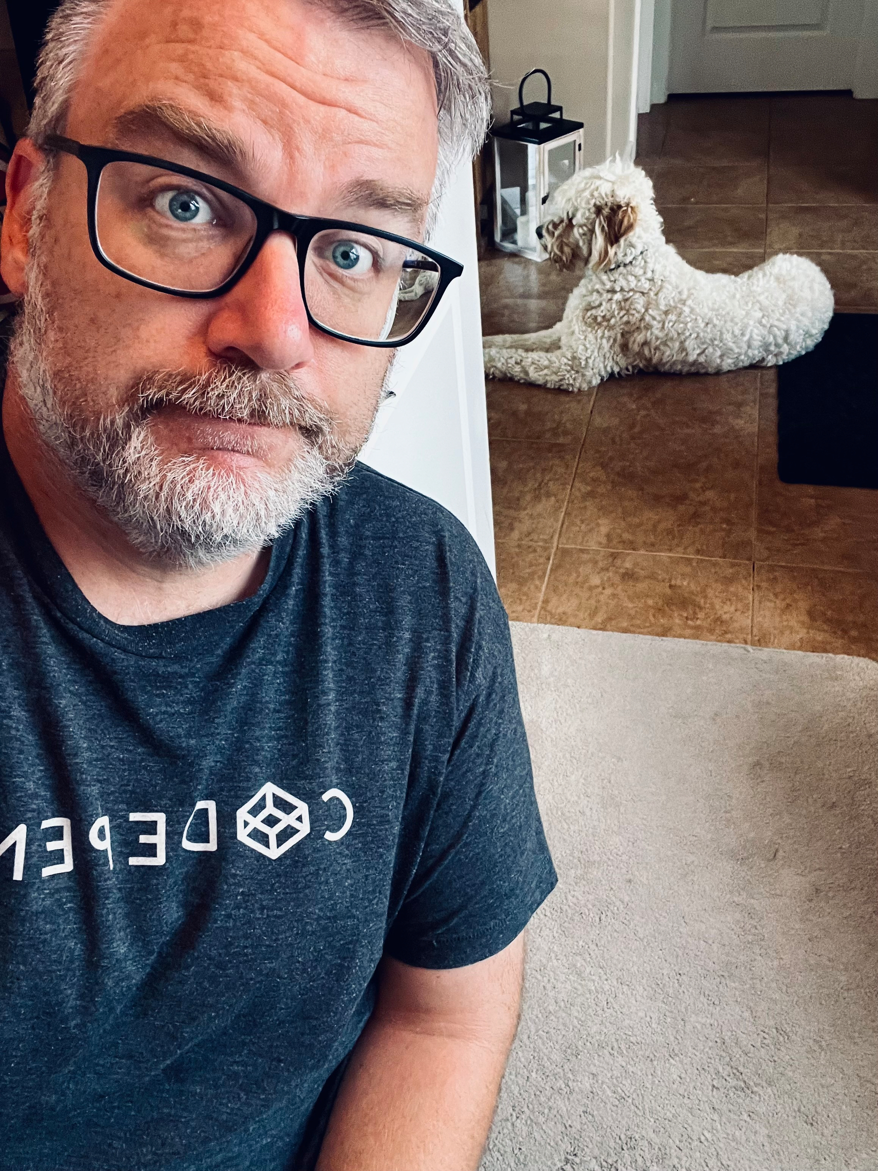 A man wearing glasses and a dark t-shirt is taking a selfie indoors while a white curly-haired dog lies on the tiled floor in the background.