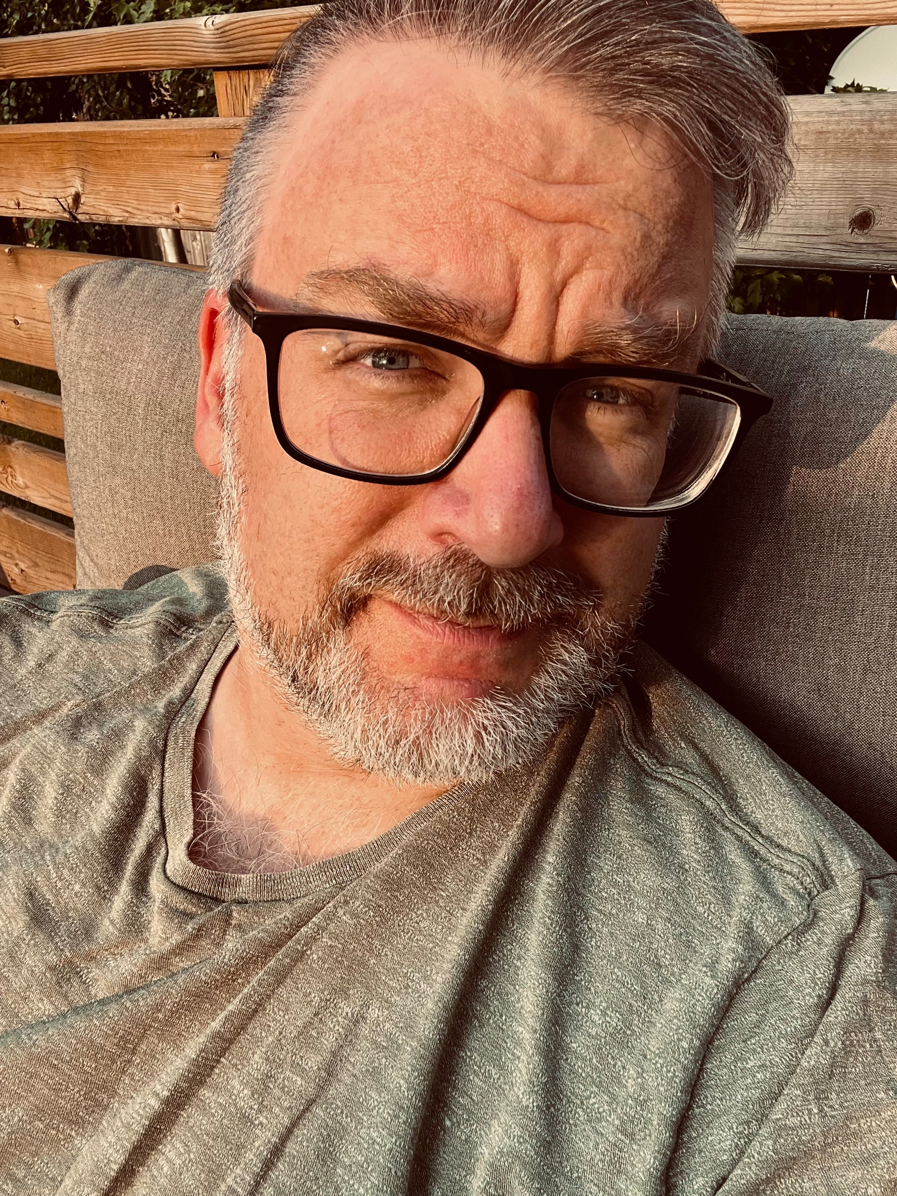 A man with gray hair, beard, and glasses is sitting on a cushioned outdoor bench, looking directly at the camera.