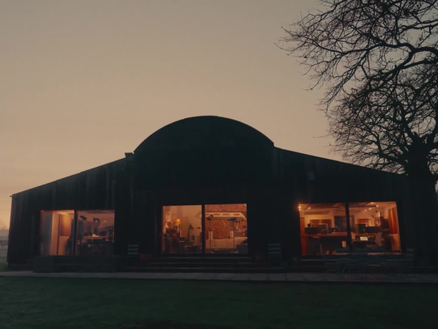 A large, rustic building with illuminated interior spaces is set against a dusky sky and framed by a tree on the right.