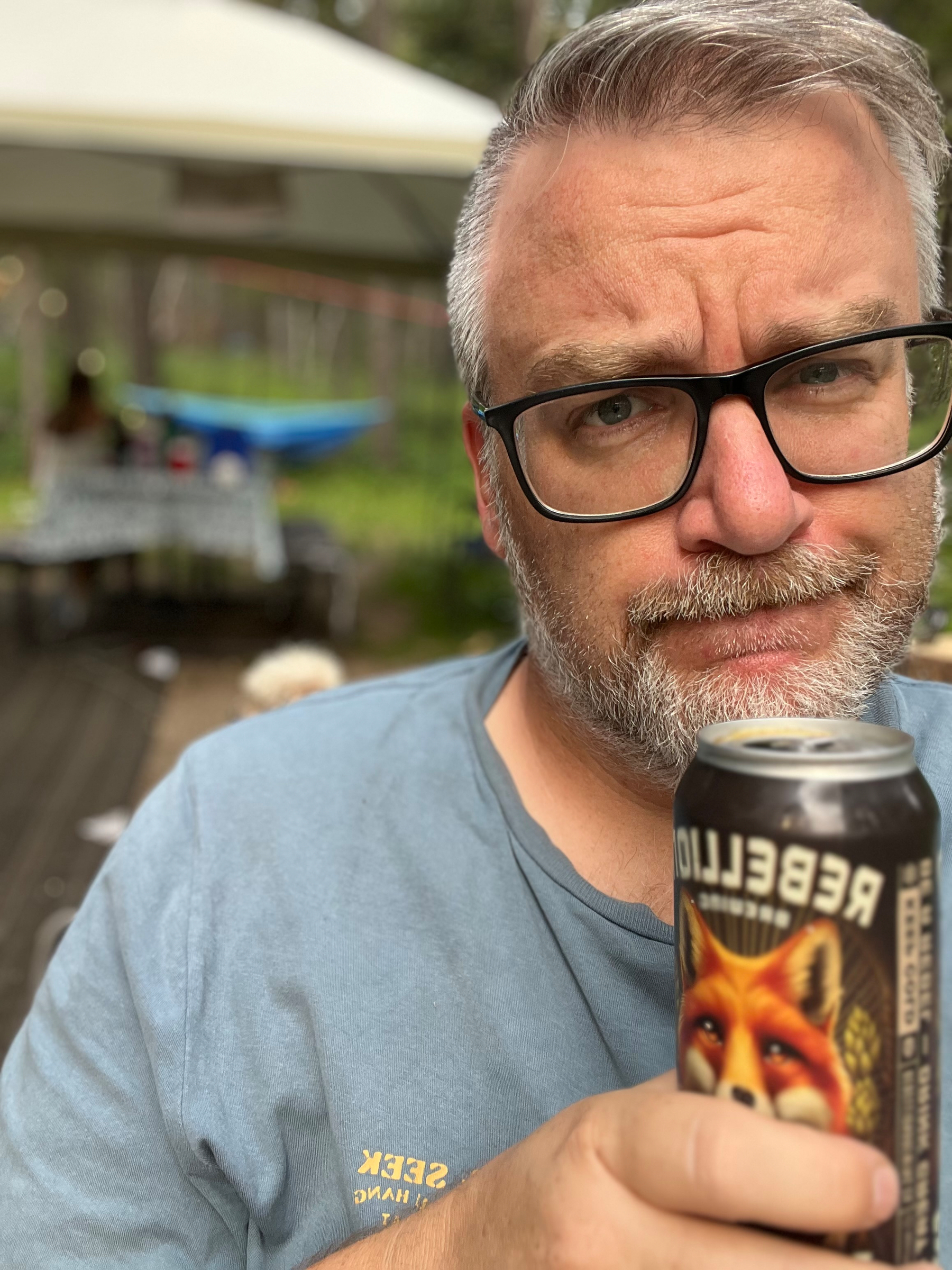 A man with glasses and a beard is holding a can of Rebellion beer and looking at the camera with a slightly raised eyebrow.