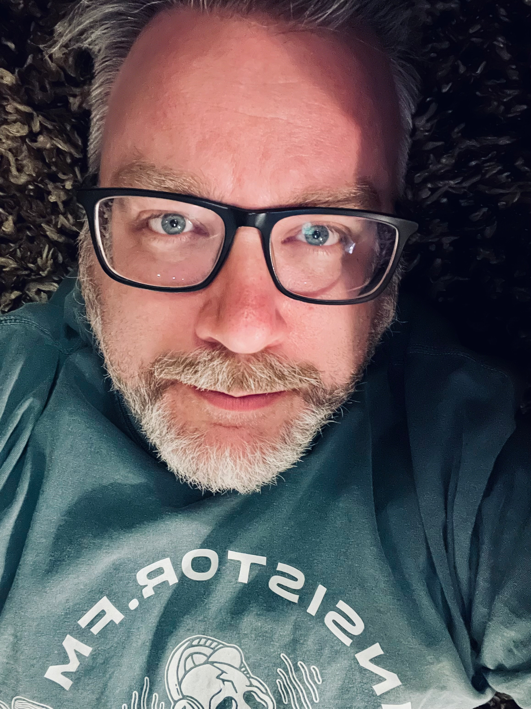 A person with glasses and a beard is lying down, wearing a dark shirt with a graphic design.