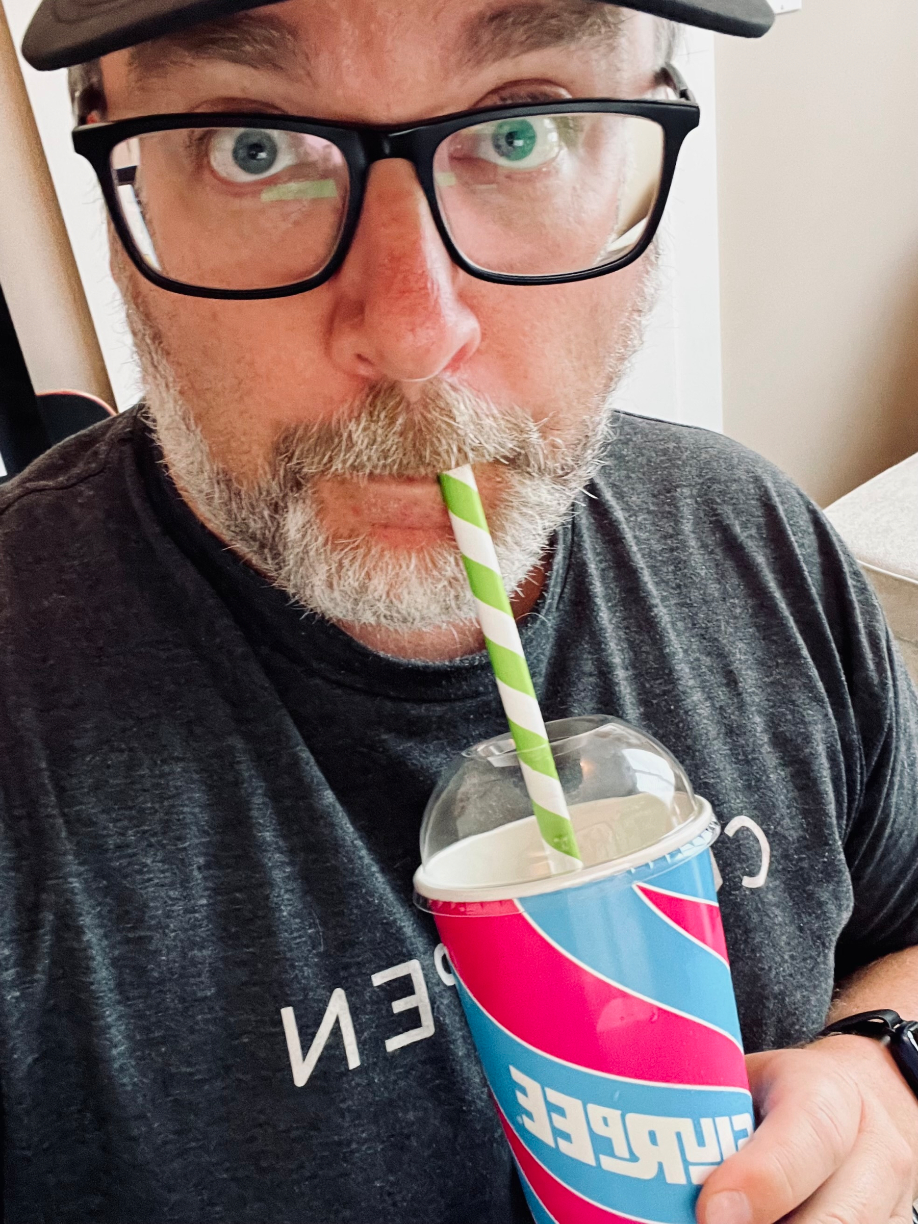 A man with glasses and a beard is sipping a colorful Slurpee through a striped straw.