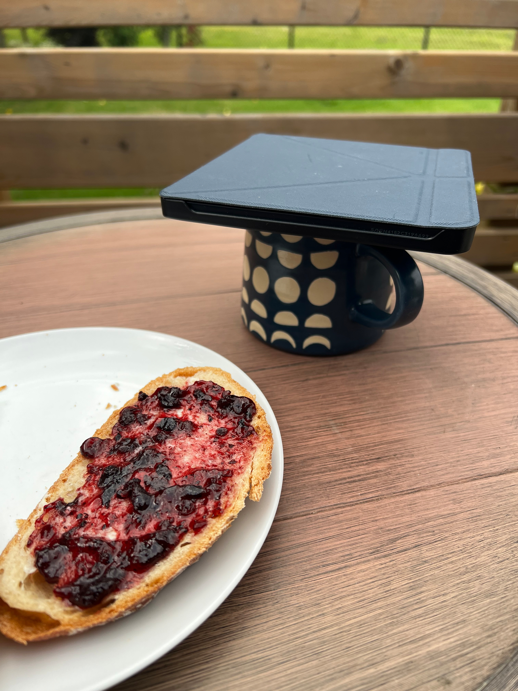 A slice of bread with berry jam on a white plate rests on a wooden table next to a black mug with a geometric pattern, topped with a closed Kobo tablet. 