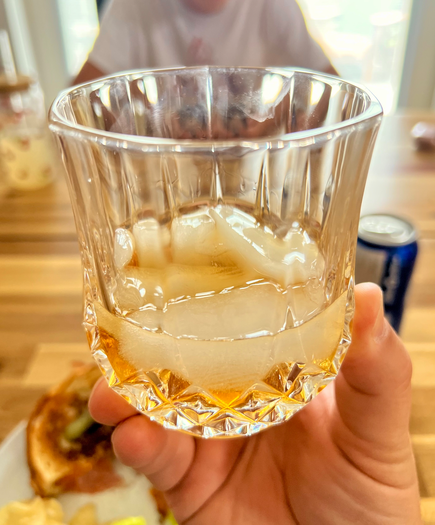 A hand is holding a glass with ice and a small amount of amber-colored liquid, with a blurred background showing food and a person.