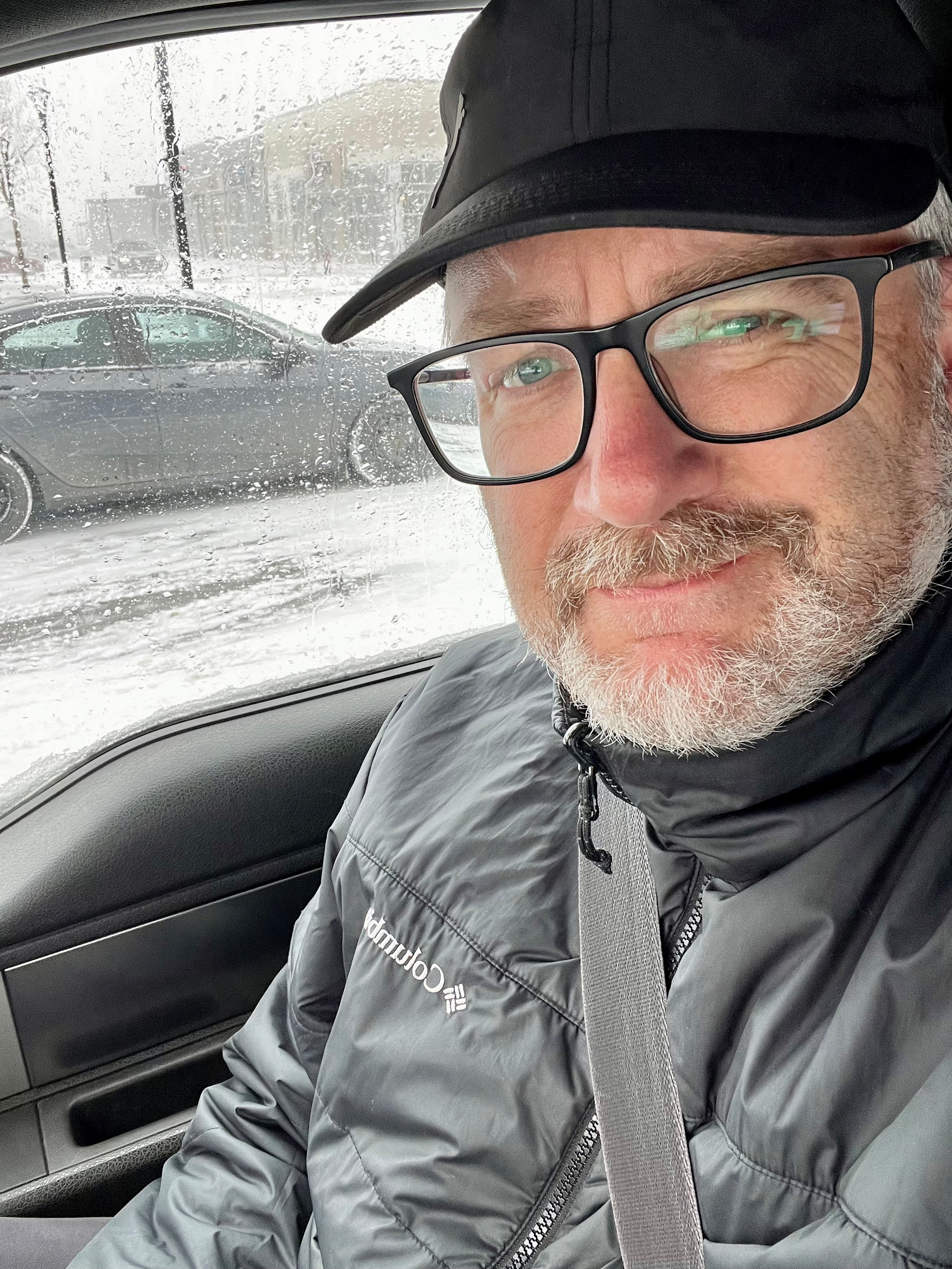 A man wearing a dark jacket and cap is sitting in a car on a snowy day with another vehicle visible through the window.