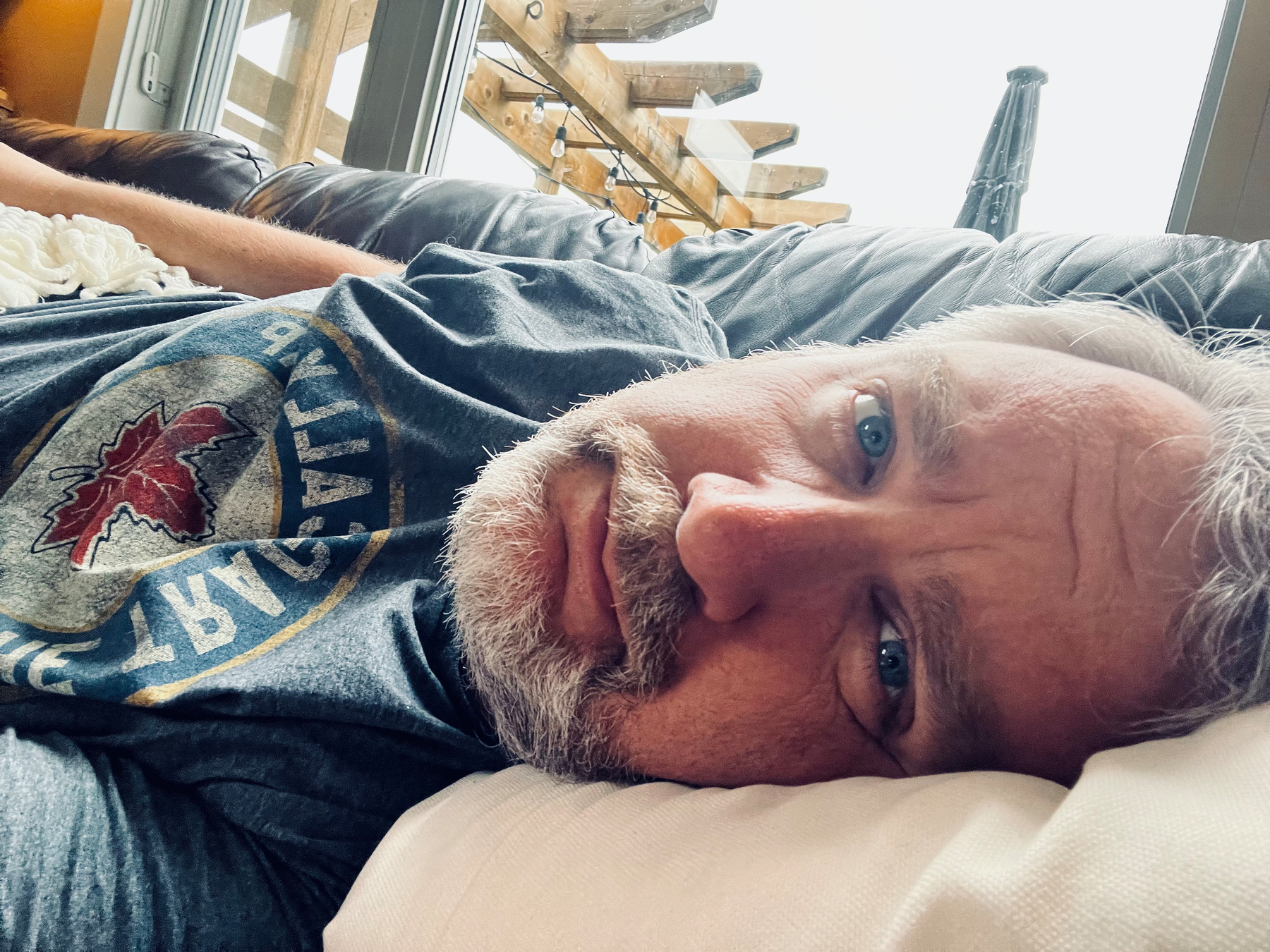 A man with light gray hair and a beard is lying on a couch, resting his head on a pillow and looking toward the camera.