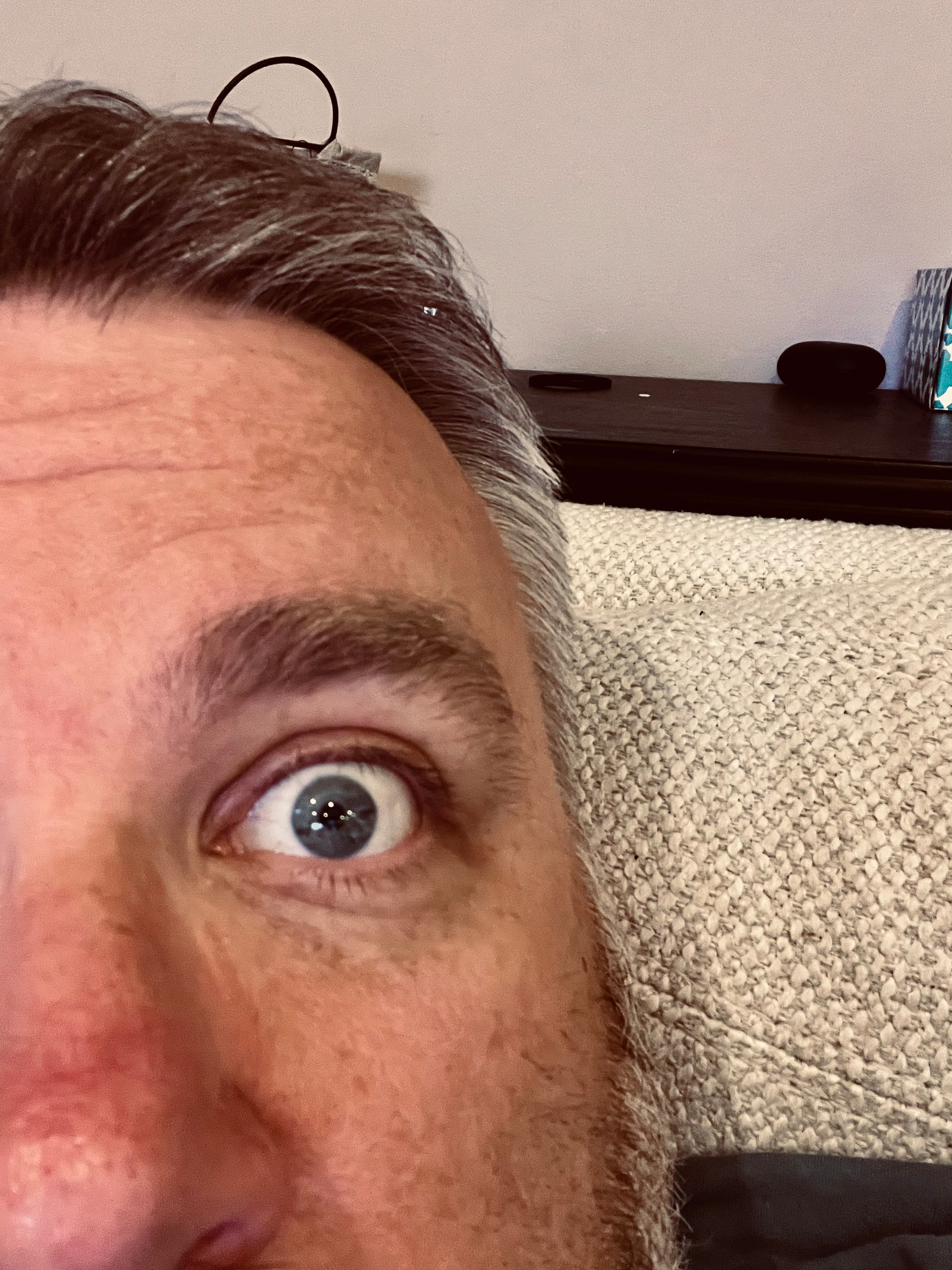 A close-up of a person's face with an eye wide open, partially lying on a textured surface, with a shelf and objects in the background.