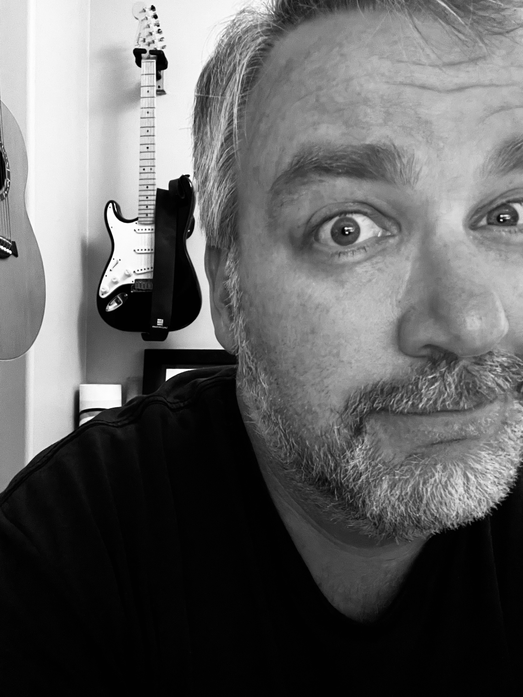 A bearded person is partially visible in the foreground, with two guitars hanging on the wall behind them.