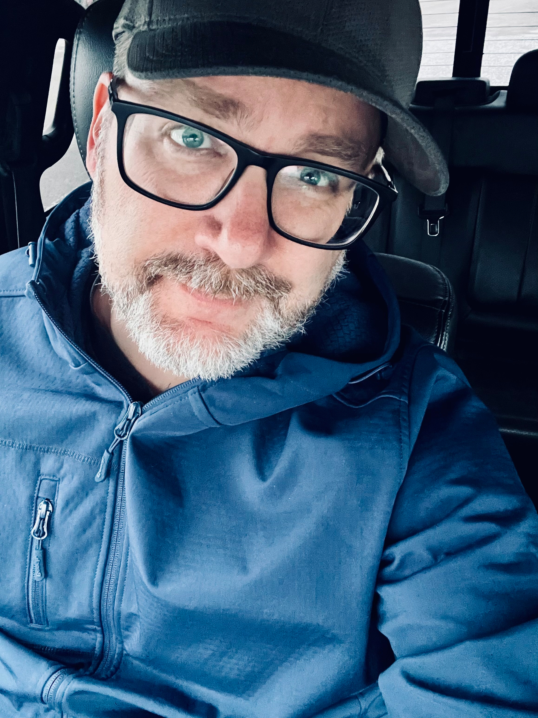 A person wearing glasses, a cap, and a blue jacket poses for a selfie inside a vehicle.