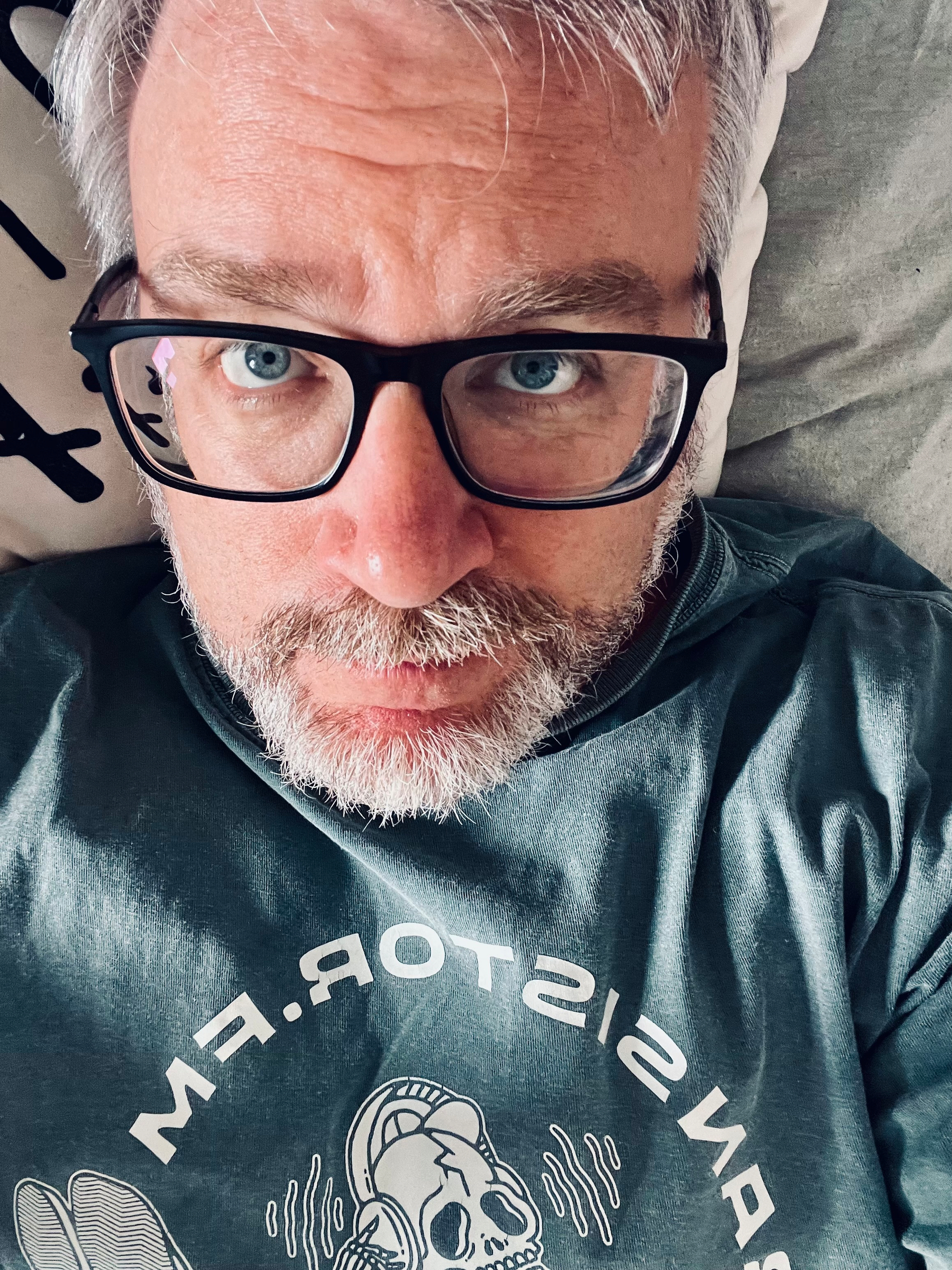 A person with glasses and a beard is lying down, looking up, wearing a shirt with a graphic design.