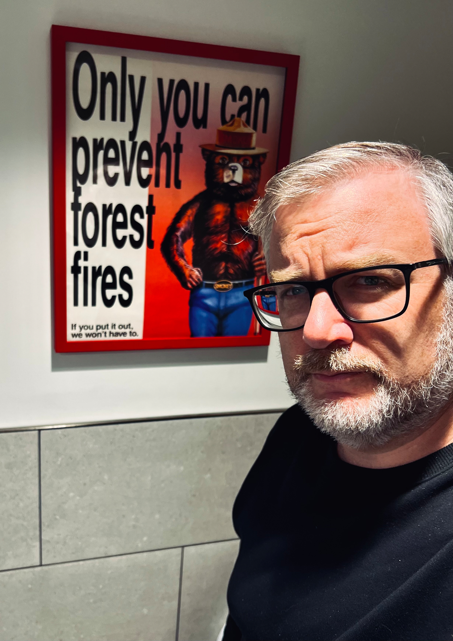 A man with glasses and gray hair stands in front of a retro-style Smokey Bear poster that says Only you can prevent forest fires.