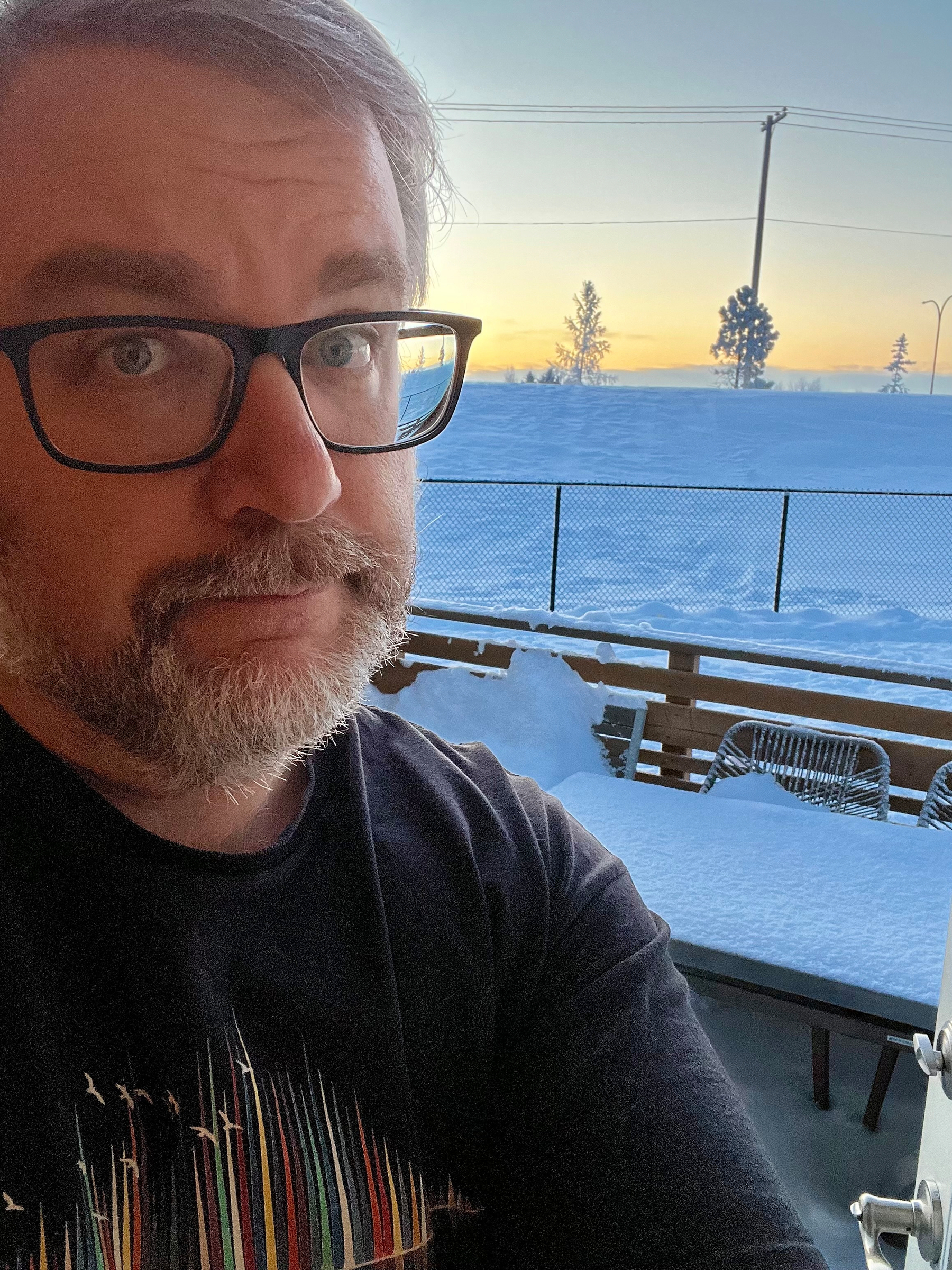A person wearing glasses stands on a snowy patio during sunset, with snow-covered furniture and a fence in the background.