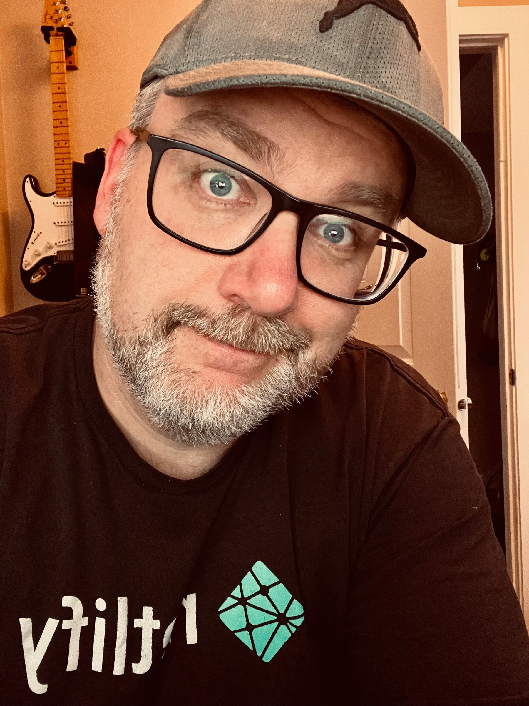 A person wearing glasses, a baseball cap, and a Netlify t-shirt is posing in a room with a guitar hanging on the wall.