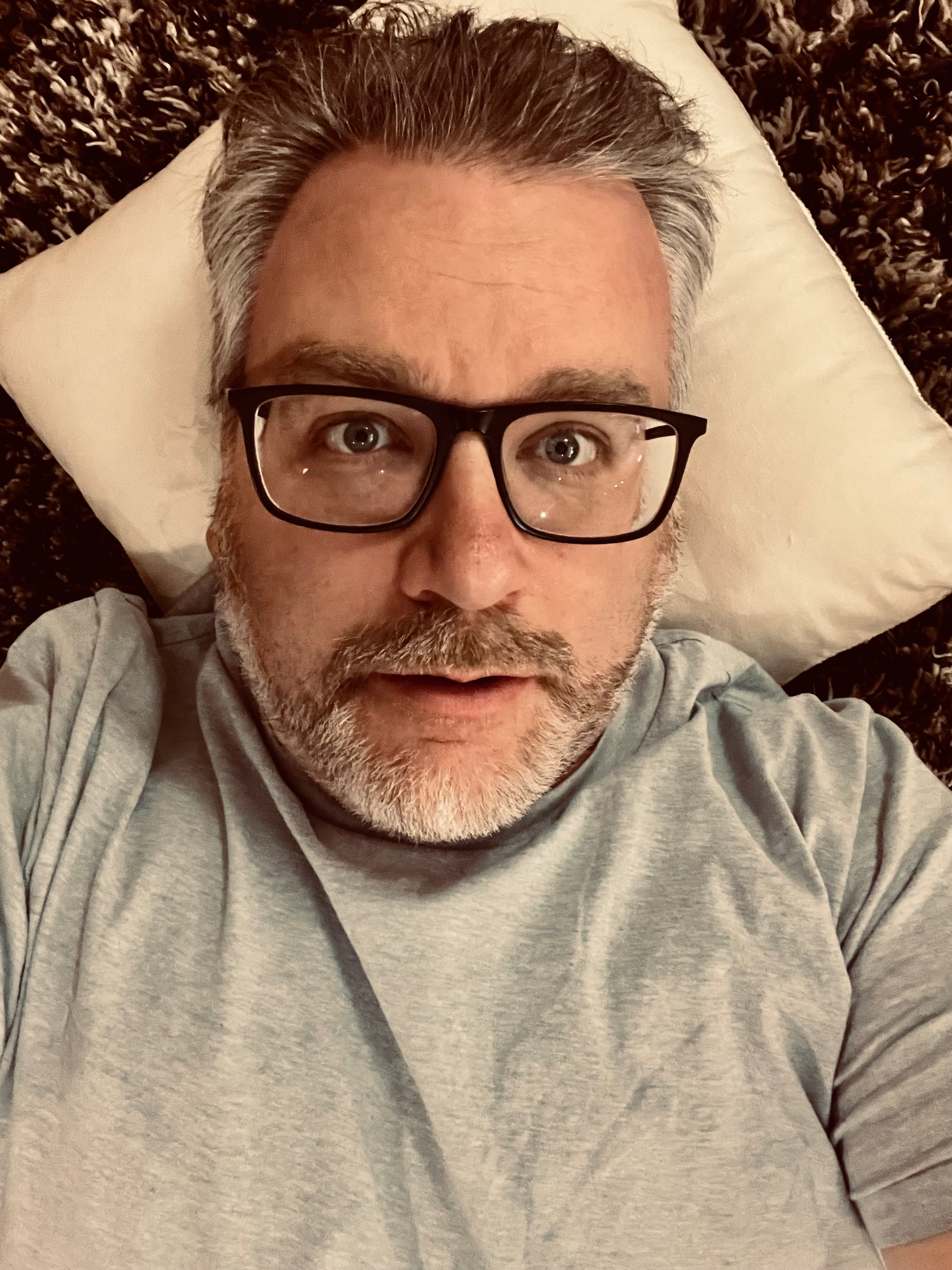 A man with glasses and a beard is lying down on a pillow, looking up.