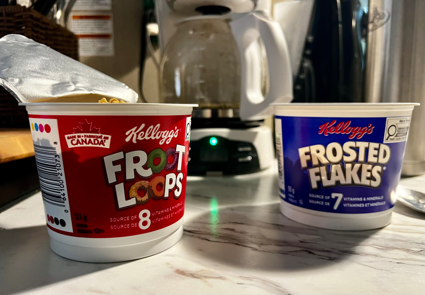 Two small Kellogg's cereal containers, one with Froot Loops and the other with Frosted Flakes, are placed on a kitchen counter with a coffee maker in the background.