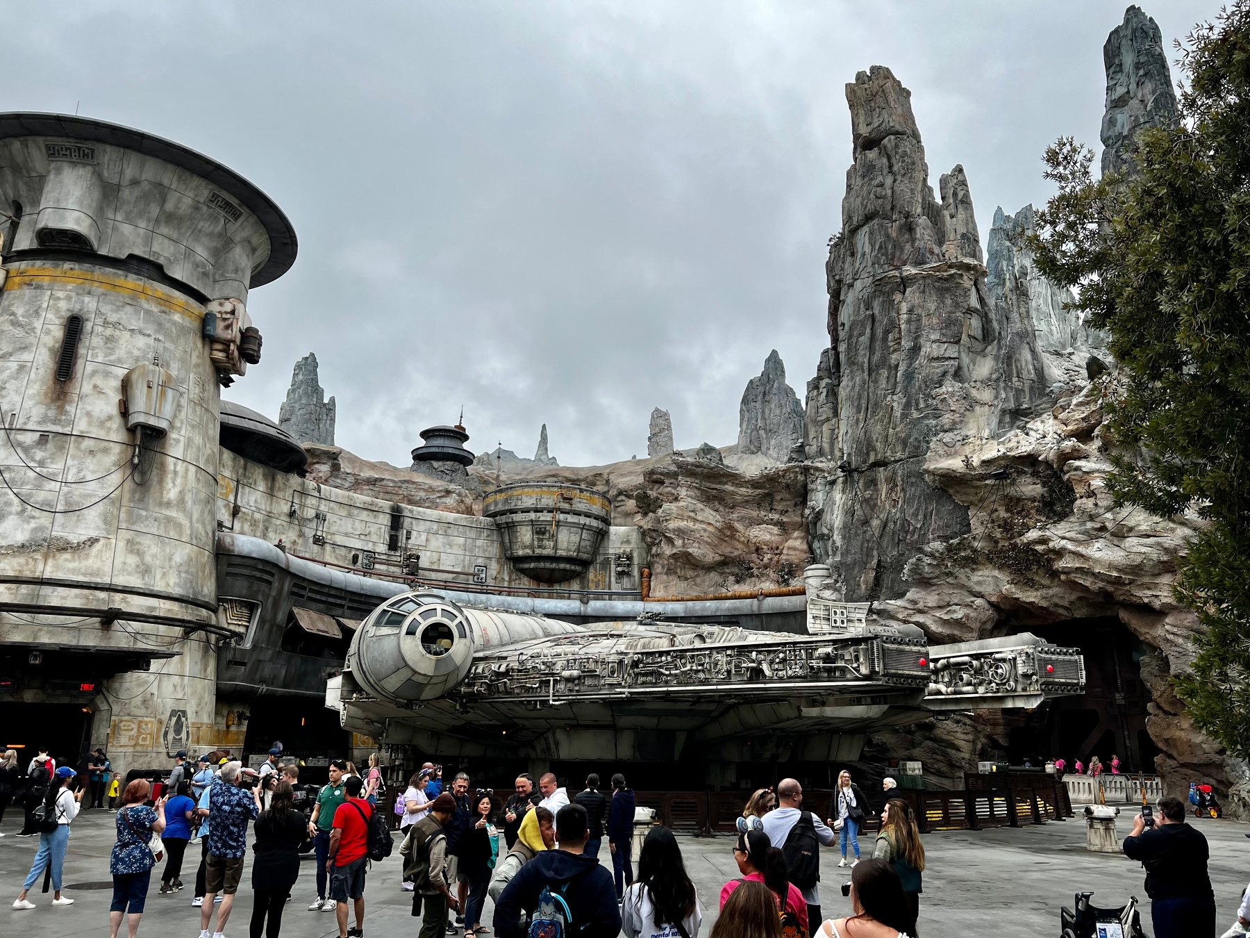 A crowd of people gathers around a life-sized Millennium Falcon replica in a detailed Star Wars-themed environment featuring rocky spires and futuristic structures. Disneyland Galaxy’s Edge 