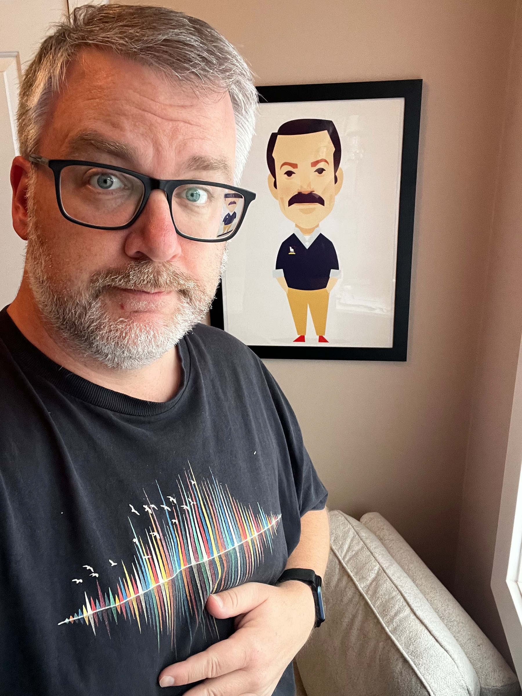A man wearing glasses and a black t-shirt with a colorful design stands beside a framed illustration of Ted Lasso on the wall.