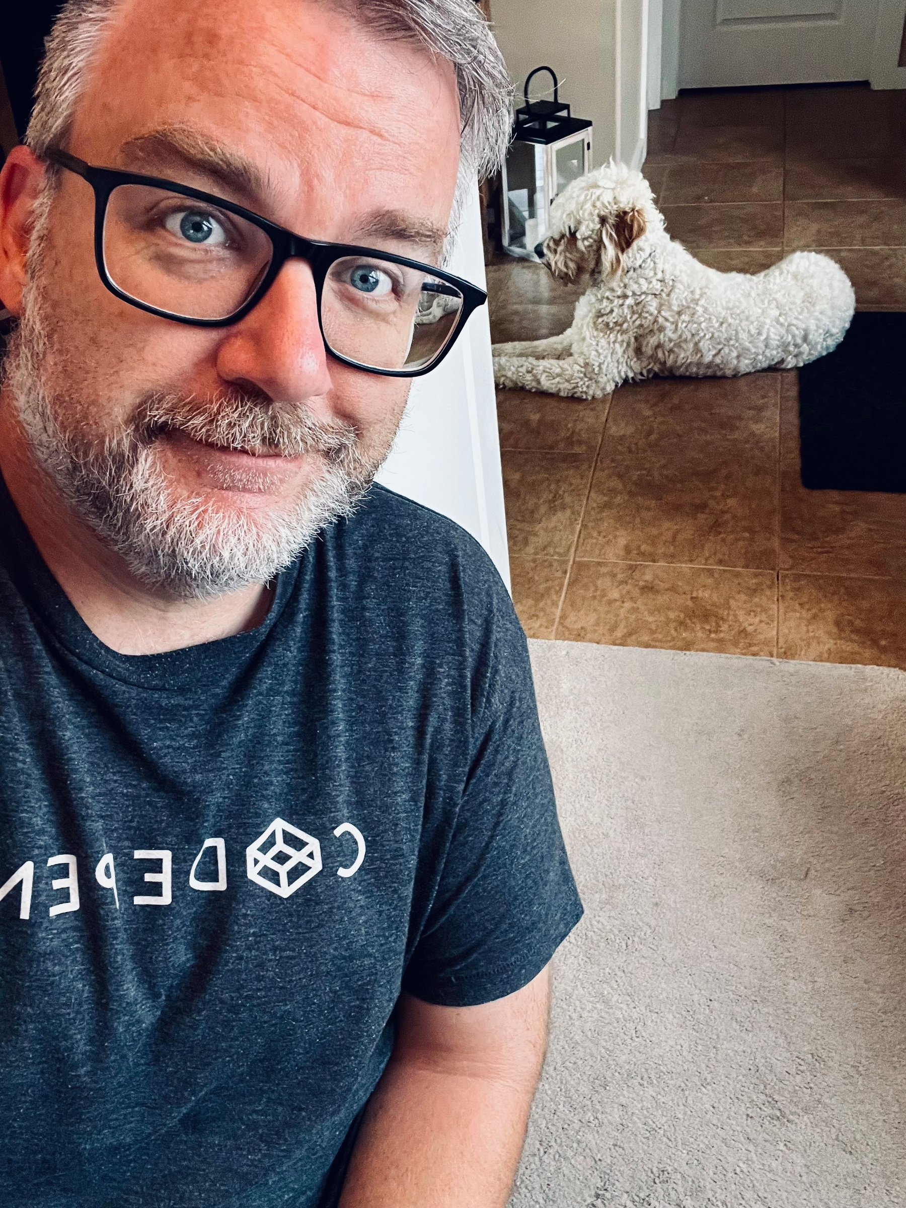 A man with glasses and a gray beard is smiling at the camera while a curly-haired white dog lies on the tiled floor in the background.