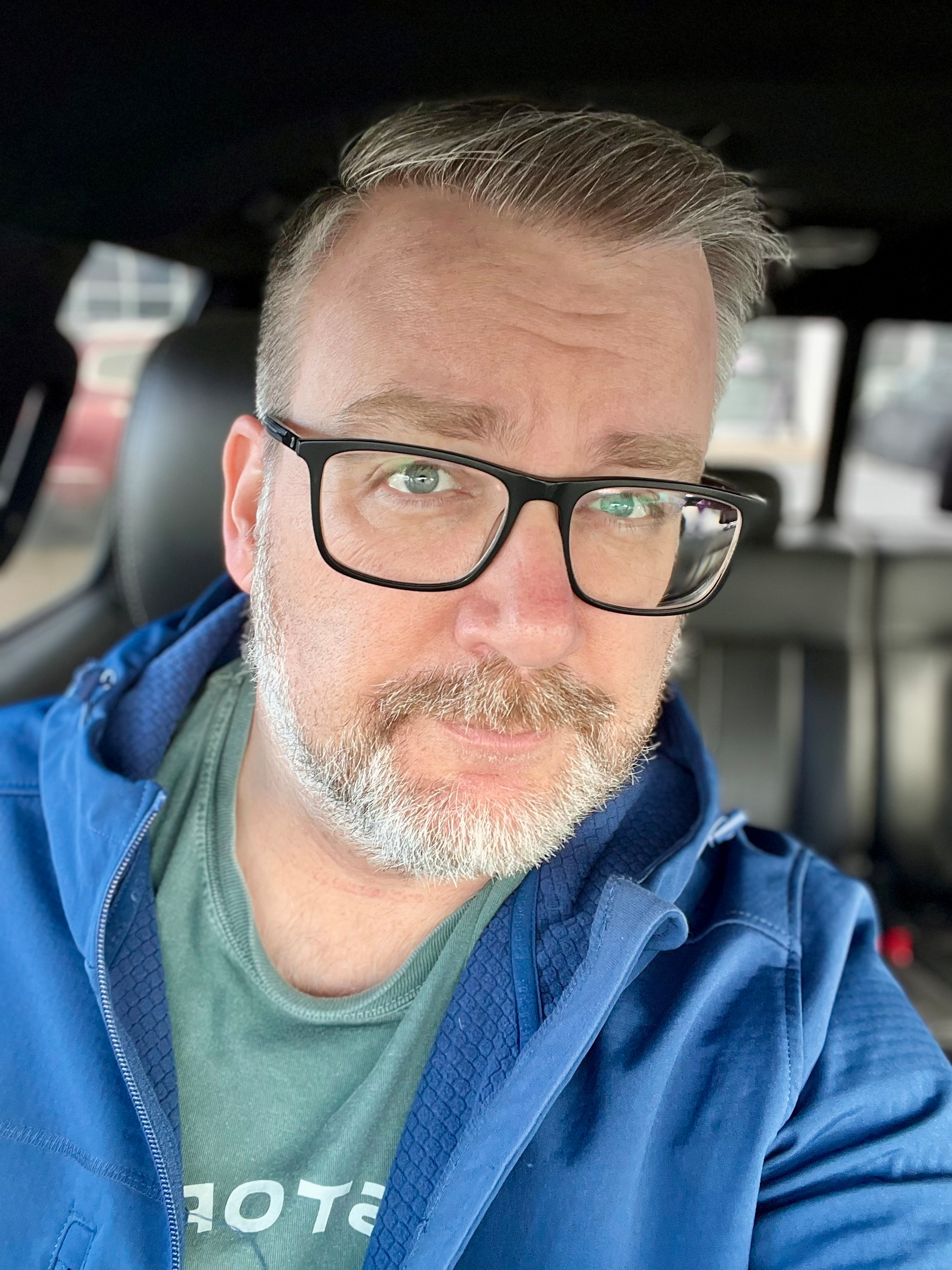A person with glasses and a beard is seated in a vehicle, wearing a blue jacket and a green shirt.