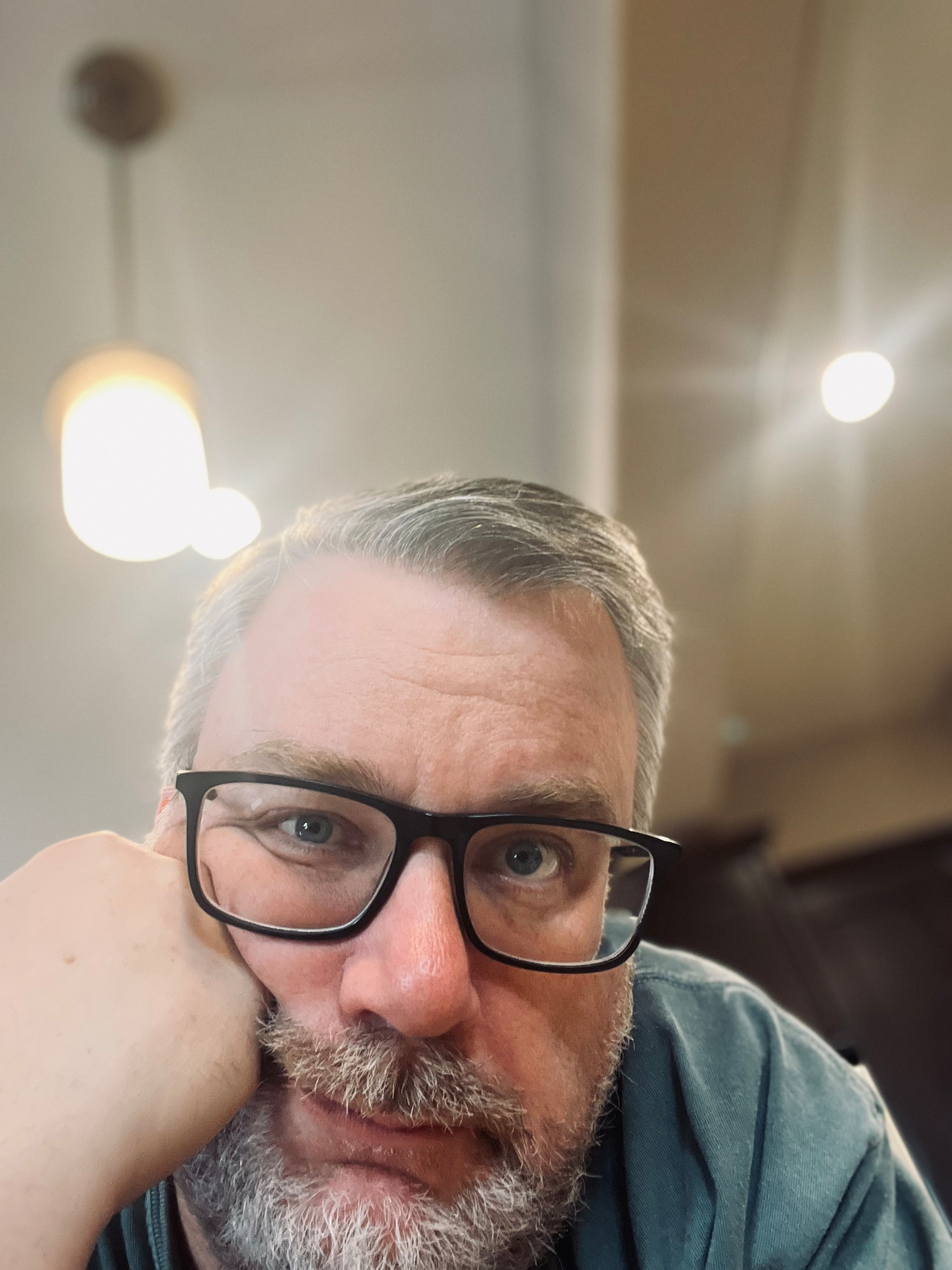 A bearded person with glasses is resting their chin on their hand, looking at the camera, with a warm indoor light in the background.