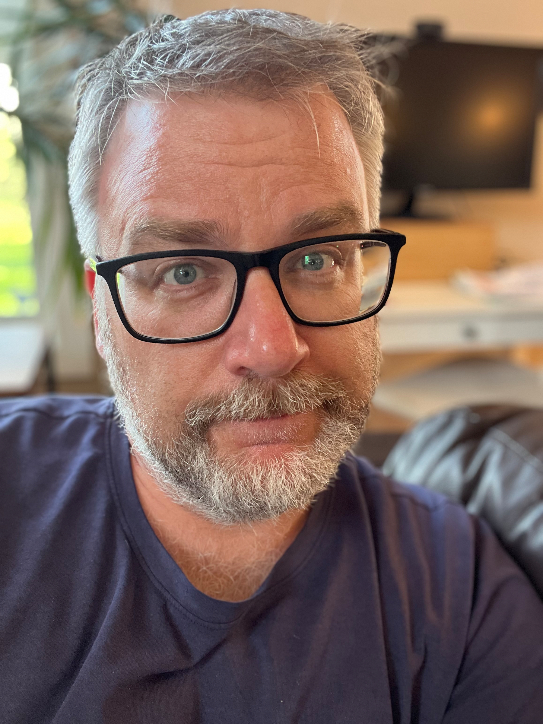 A man with glasses and a beard is sitting indoors, looking at the camera with a neutral expression.