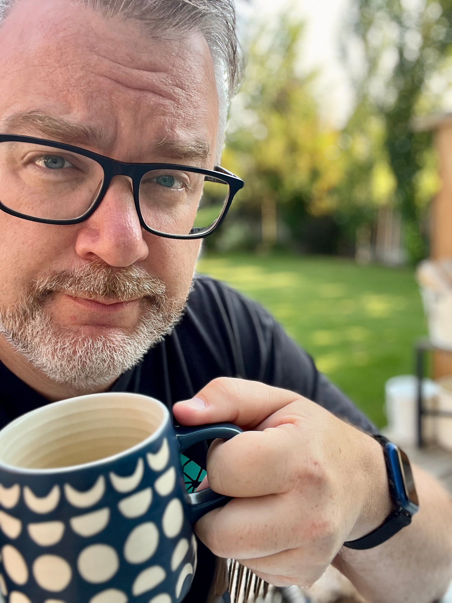 A man with glasses and a beard is holding a blue and white polka-dotted mug, looking thoughtfully at the camera while outdoors in a garden or backyard.