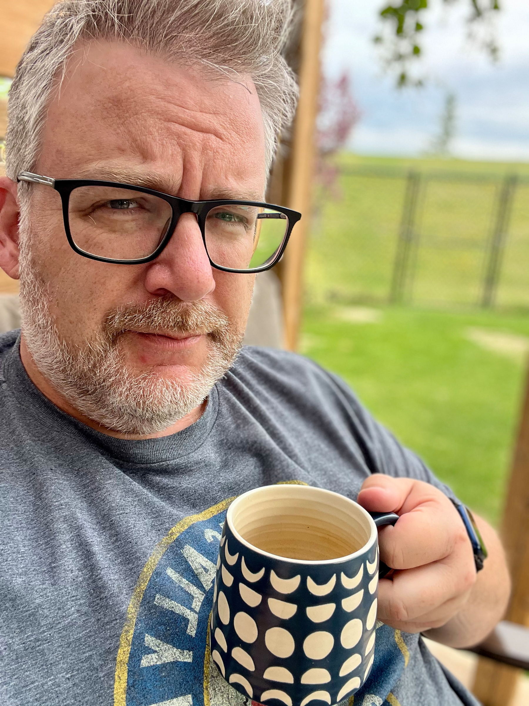 A person wearing glasses and a T-shirt is holding a polka dot mug while sitting outdoors.