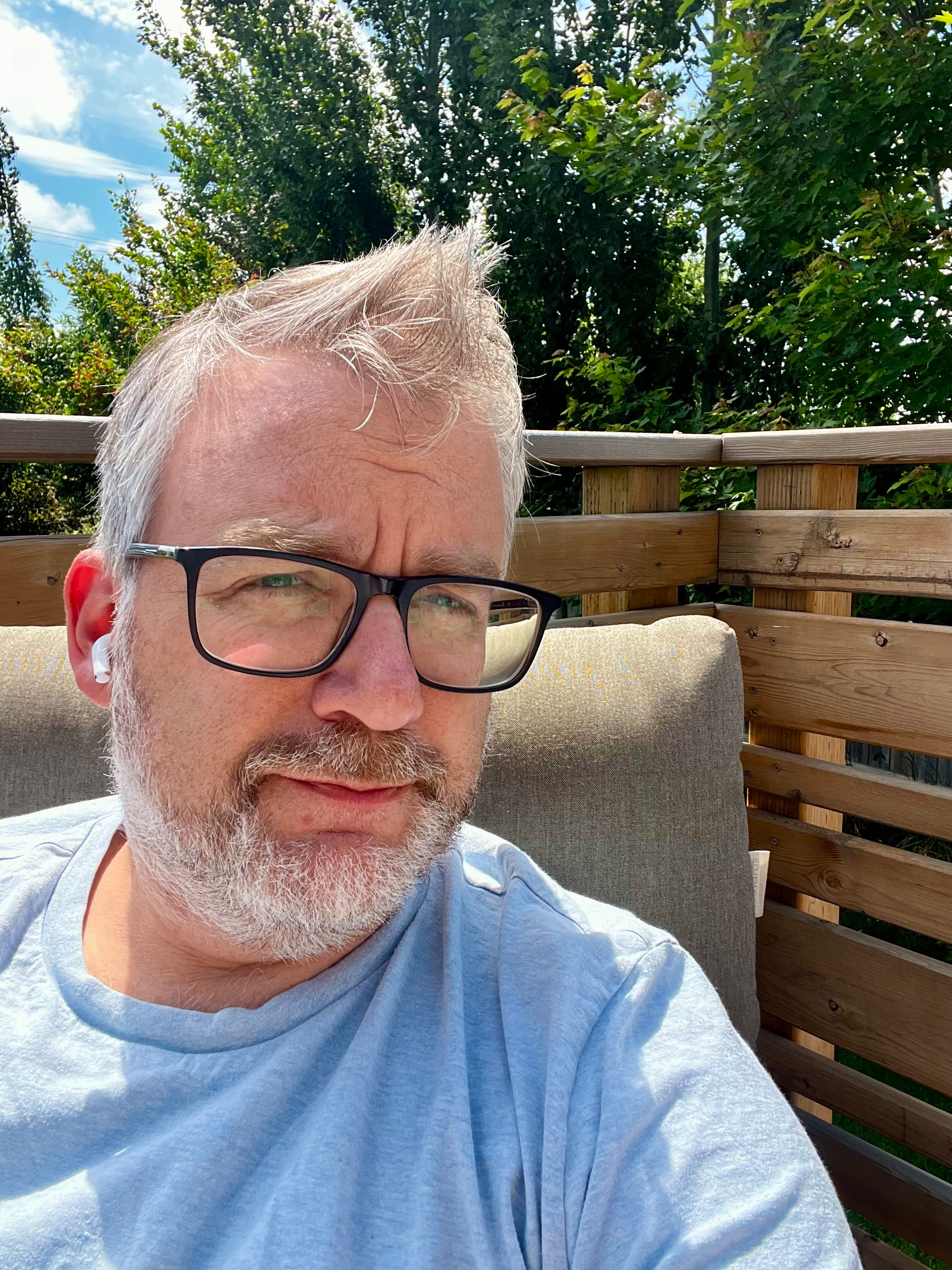 A man with glasses and a beard is sitting outdoors on a cushioned bench, surrounded by wooden fencing and greenery.