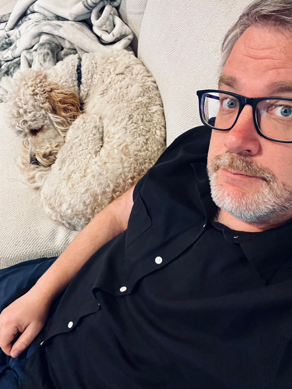 A person wearing glasses and a black shirt is sitting on a couch next to a sleeping curly-haired dog.