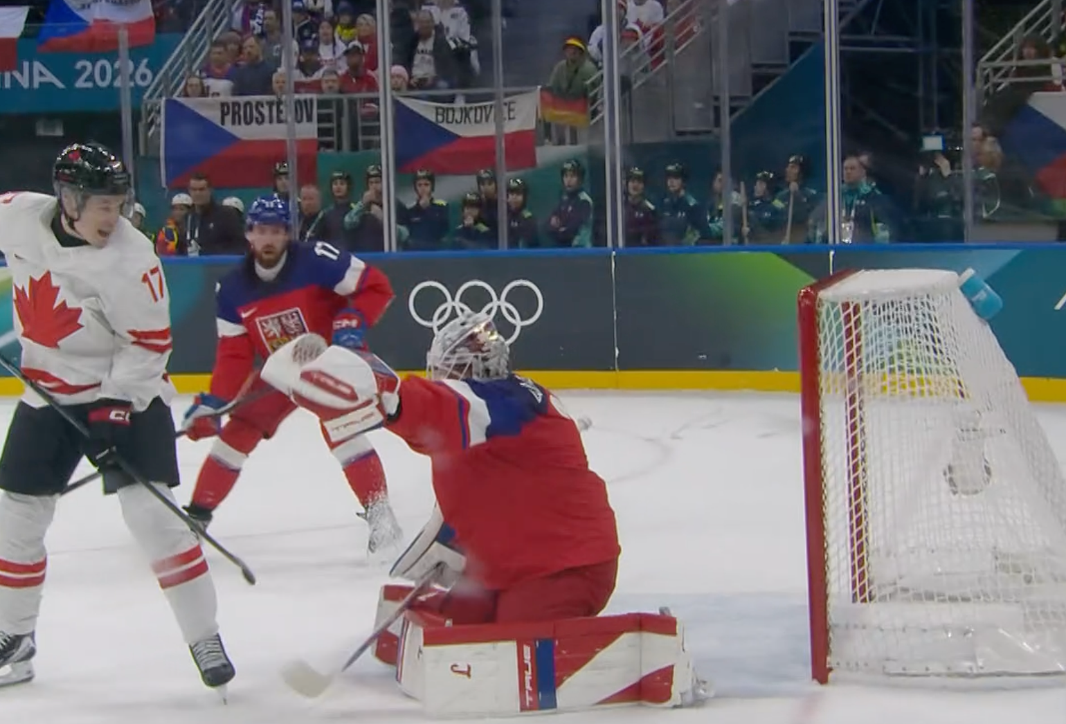 A hockey player in a red and white jersey attempts to score as the goalie, wearing red and blue, defends the net during an Olympic game.