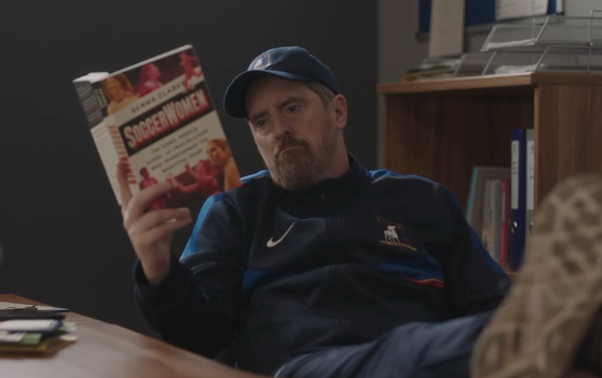 A Coach Beard from Ted Lasso wearing a cap and jacket is sitting with their feet up, reading a book about soccer women.