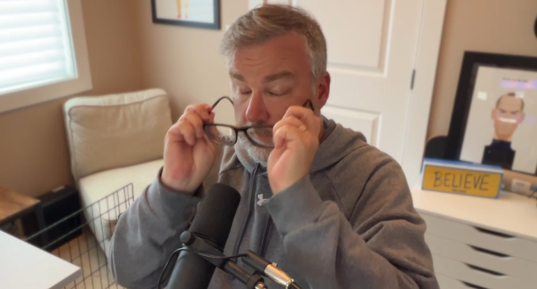 A man wearing a gray hoodie is removing his glasses while sitting in front of a microphone in a room with framed artwork and a BELIEVE sign.
