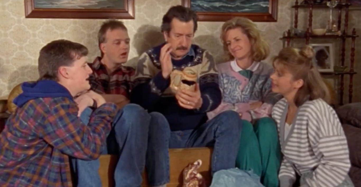 Screenshot from an Australian movie from the late ‘90s showing a family sitting around on a couch together with 3 kids and a mom all looking at the dad holding an object and looking at it with reverence and awe.