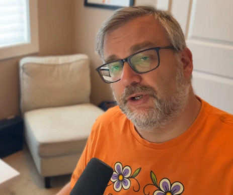 A man wearing glasses and an orange shirt with floral designs is speaking into a microphone in a room with a beige armchair and a window in the background.