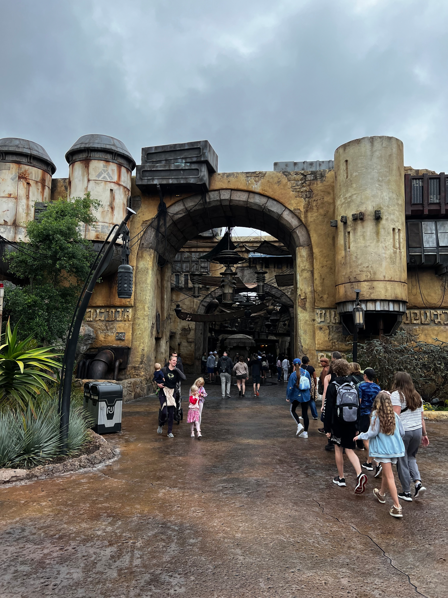 People are walking toward a weathered, sci-fi themed structure with cylindrical towers under a cloudy sky. At Disneyland Galaxy’s Edge