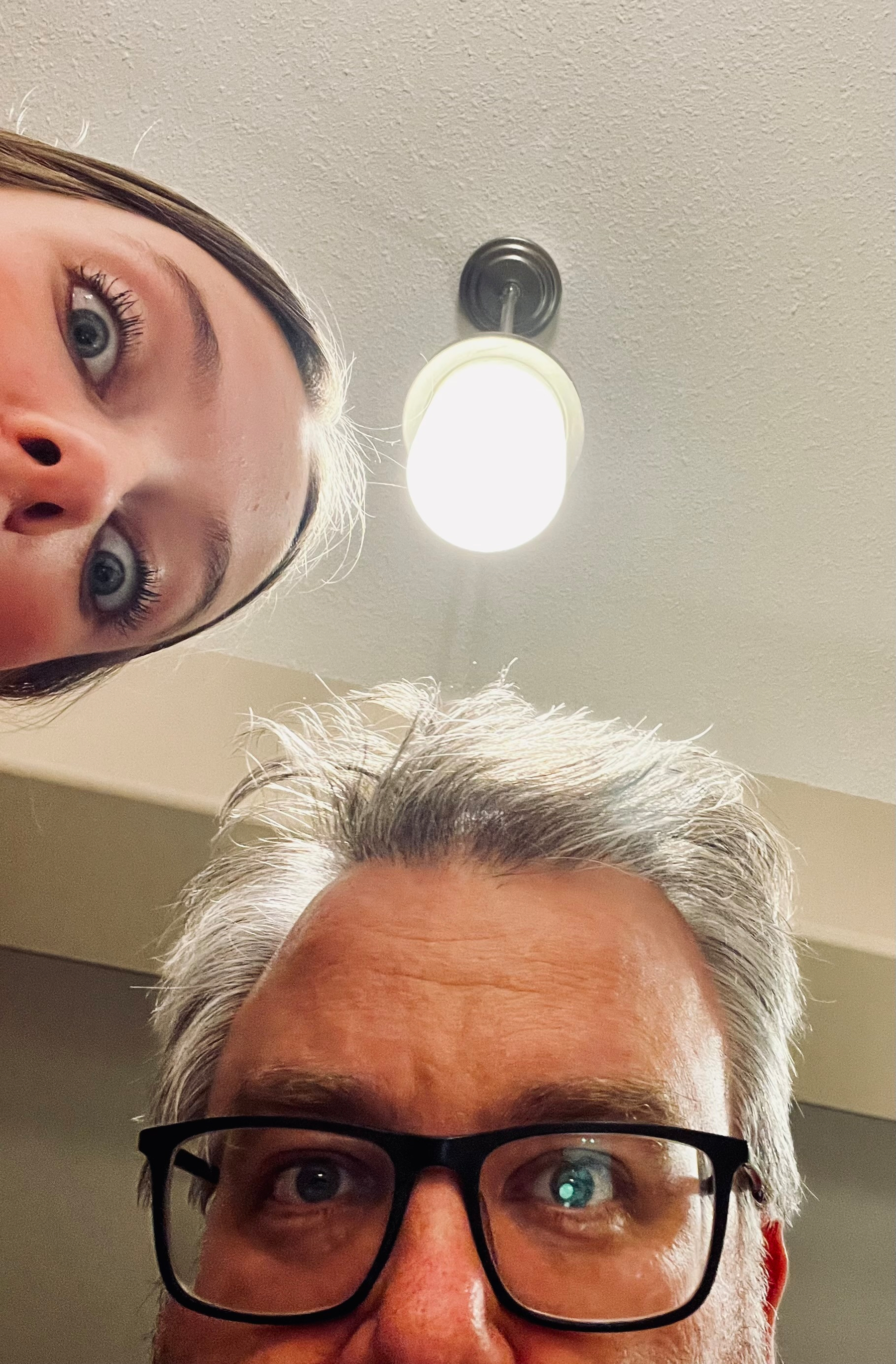 Two people with curious expressions are looking down from beneath a bright ceiling light.