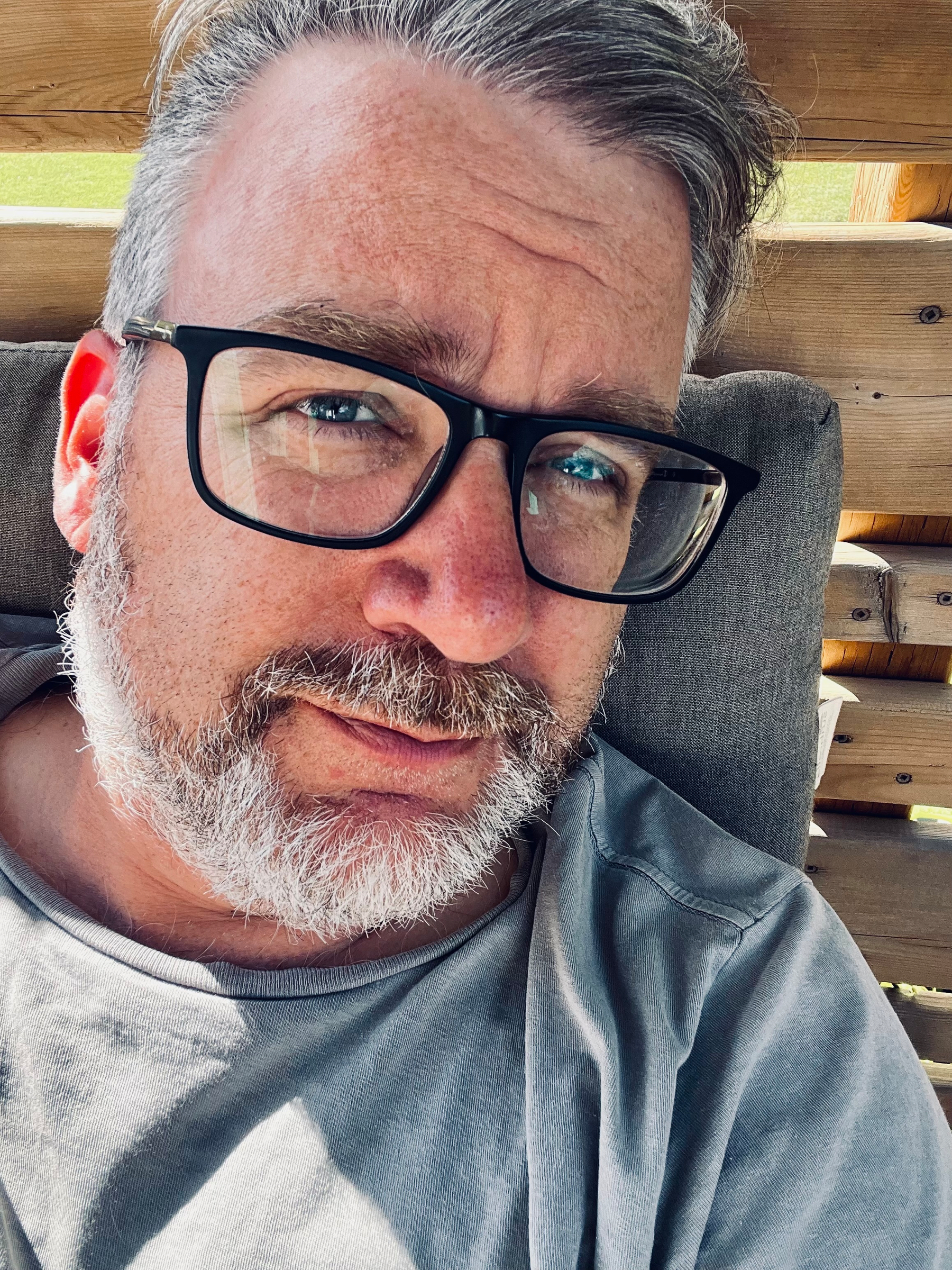 A middle-aged man with gray hair and beard is wearing black-framed glasses and a gray shirt while sitting outdoors on a cushioned wooden bench.