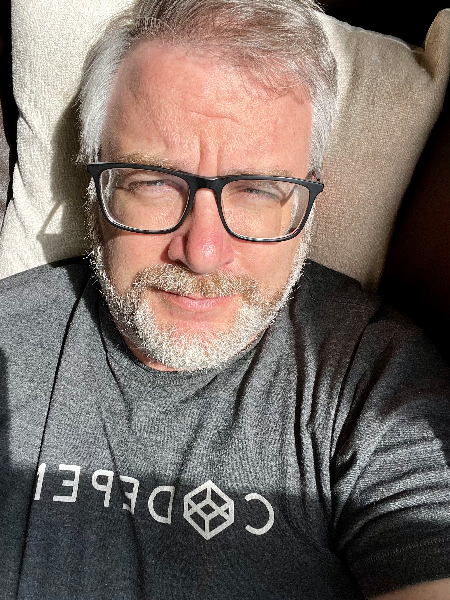 A middle-aged man with gray hair and a beard is wearing black-framed glasses and a gray t-shirt with a logo, sitting or lying on a light-colored cushion in bright natural light.