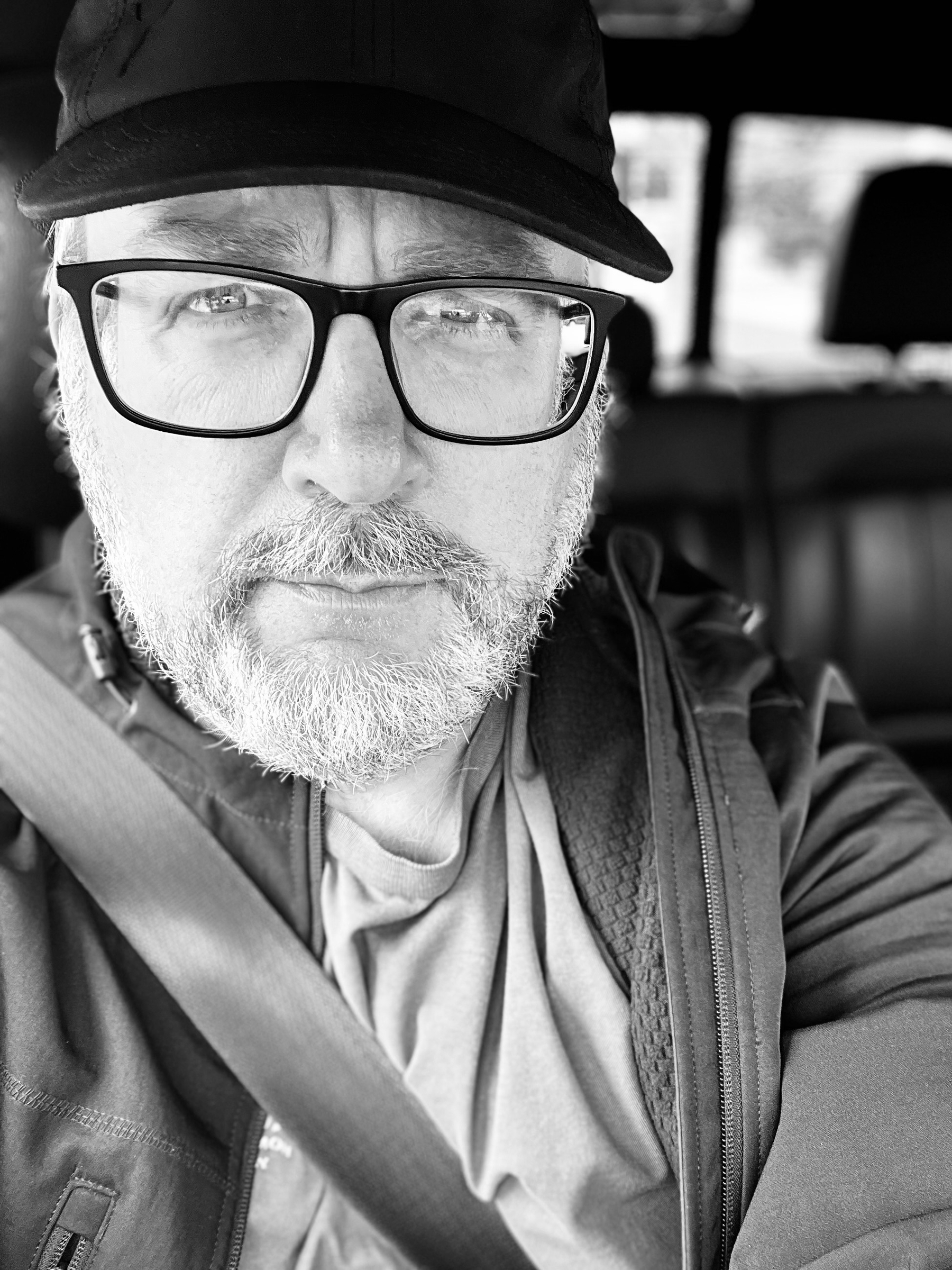 A bearded man wearing glasses and a cap is seated in a car, captured in a black-and-white selfie.