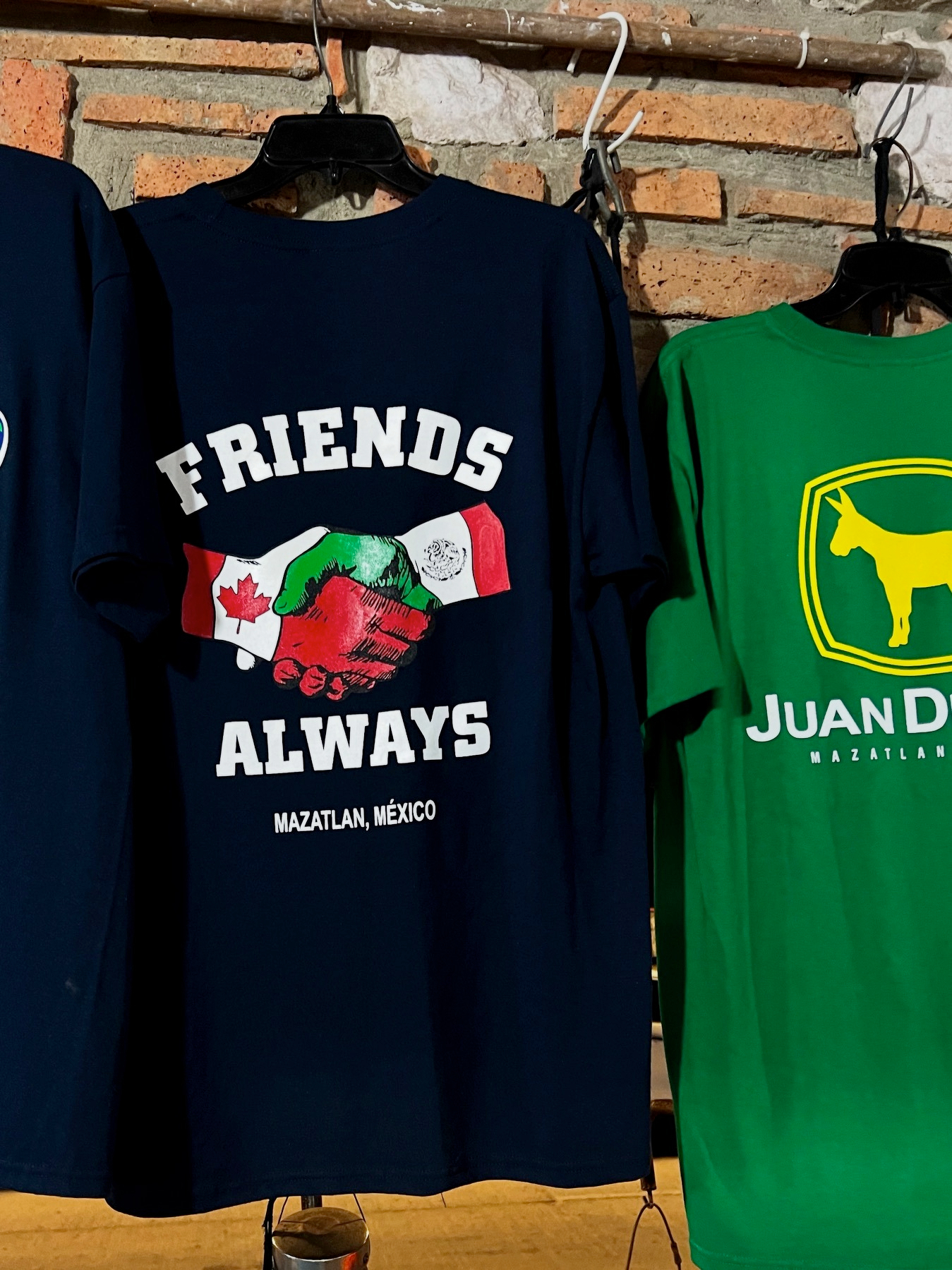 Two T-shirts are displayed, one with the text FRIENDS ALWAYS featuring clasped hands with Canadian and Mexican flags, and the other with a donkey logo and Juan Deere text.