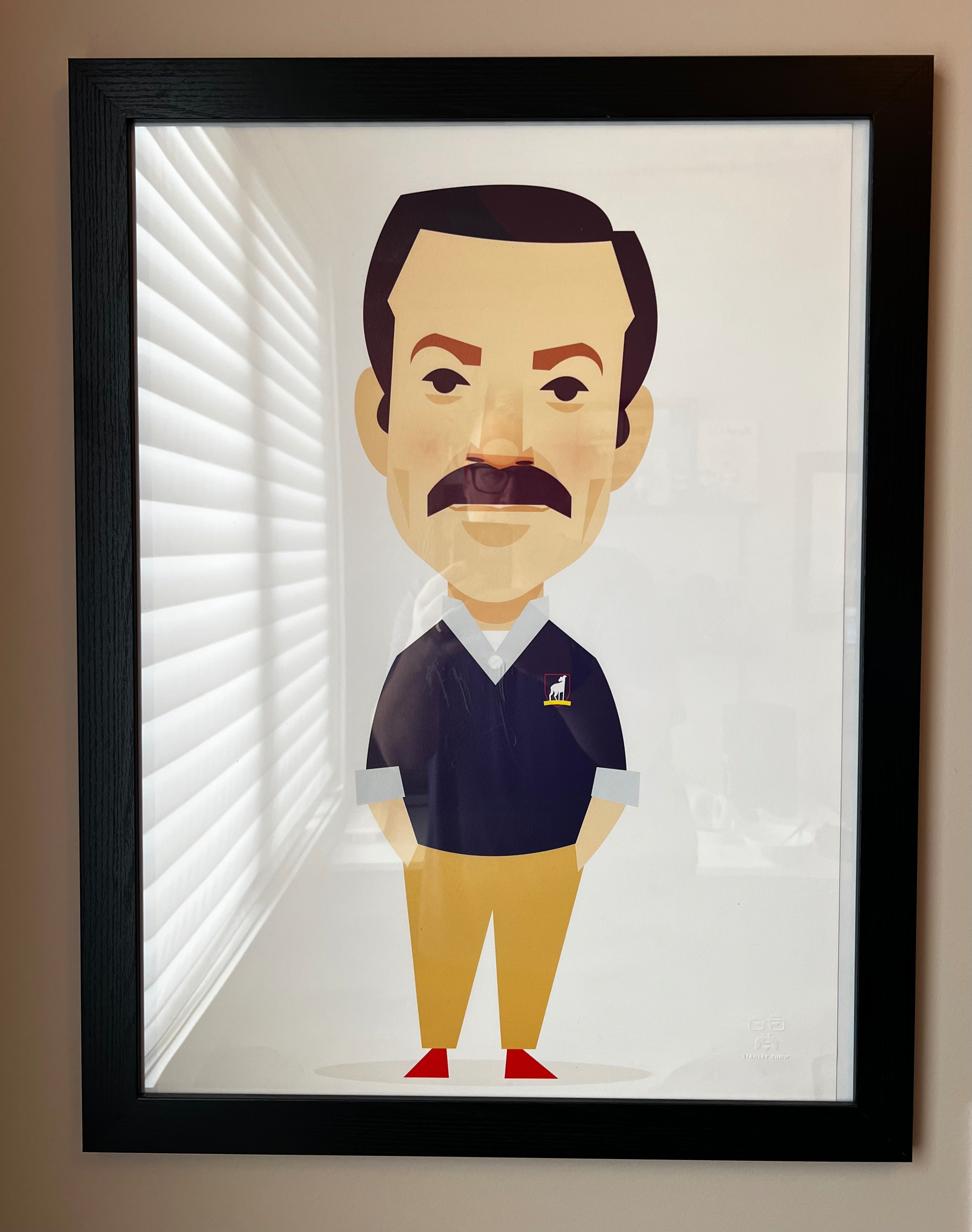A framed cartoon-style illustration of Ted Lasso with a mustache, wearing a dark sweater with a small logo, tan pants, and red shoes is displayed on a wall.