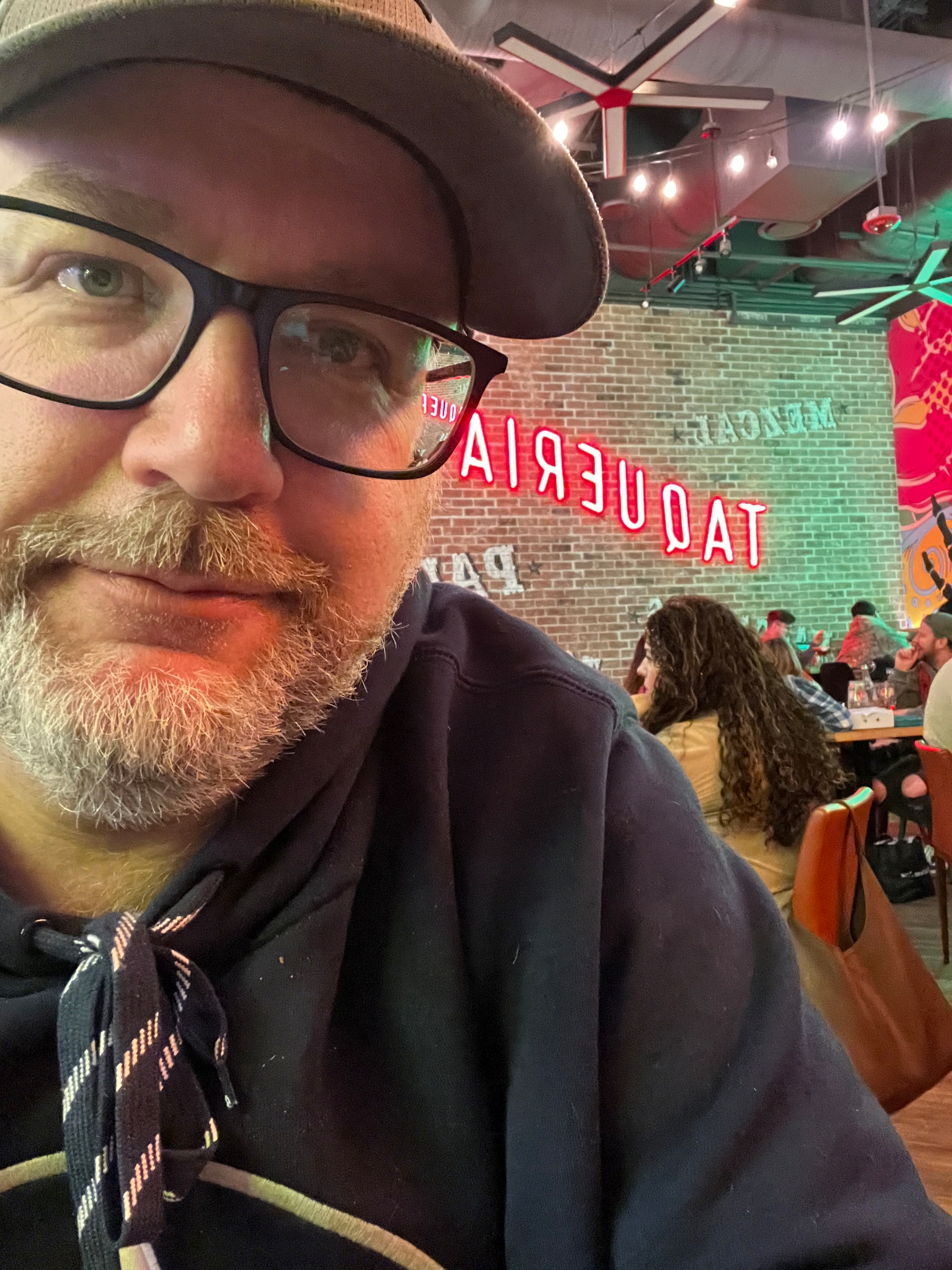 A person wearing glasses and a cap is sitting in a restaurant with a neon Taqueria sign and people dining in the background.