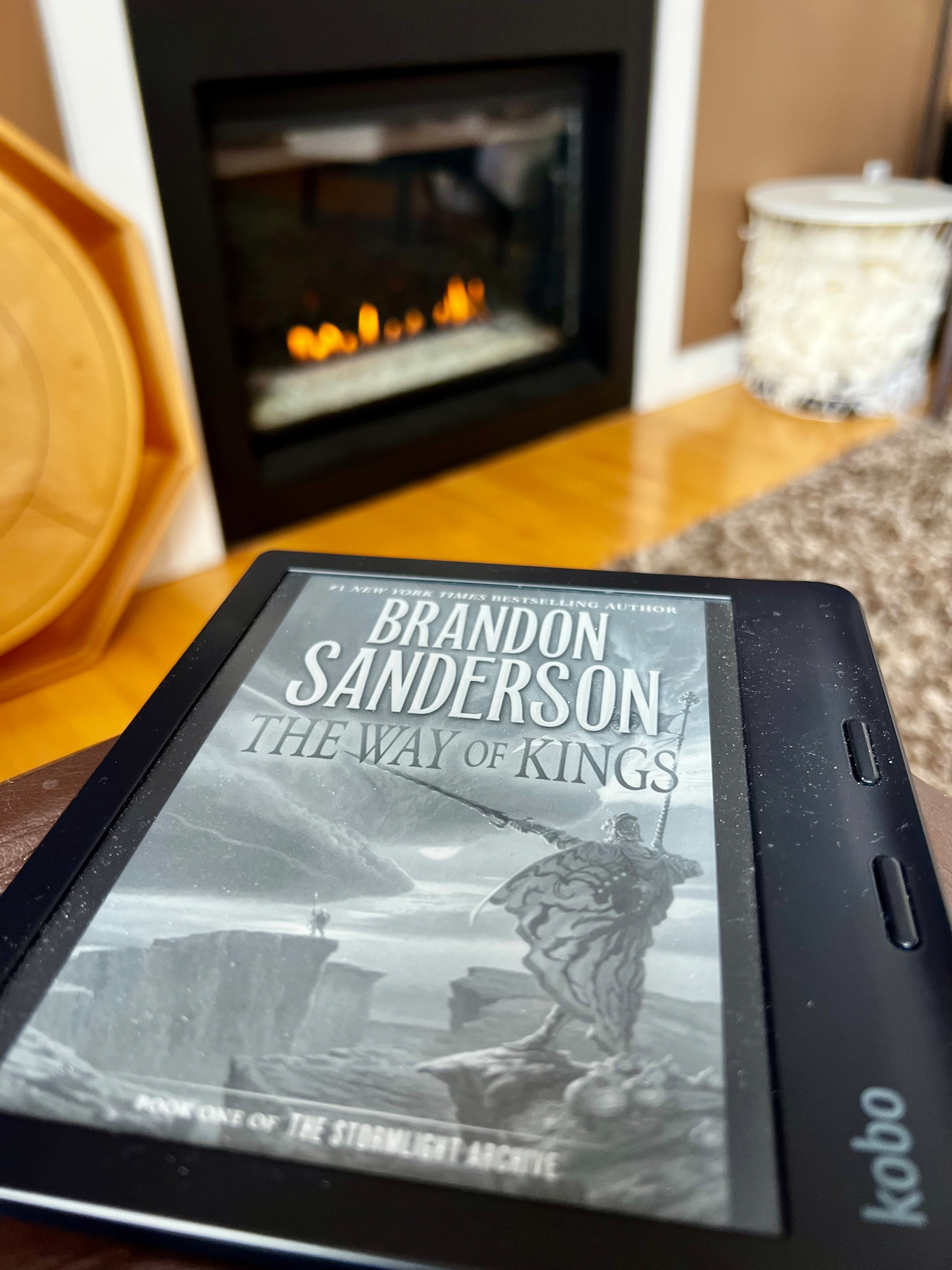 A Kobo e-reader displays the cover of The Way of Kings by Brandon Sanderson, positioned in front of a cozy fireplace.